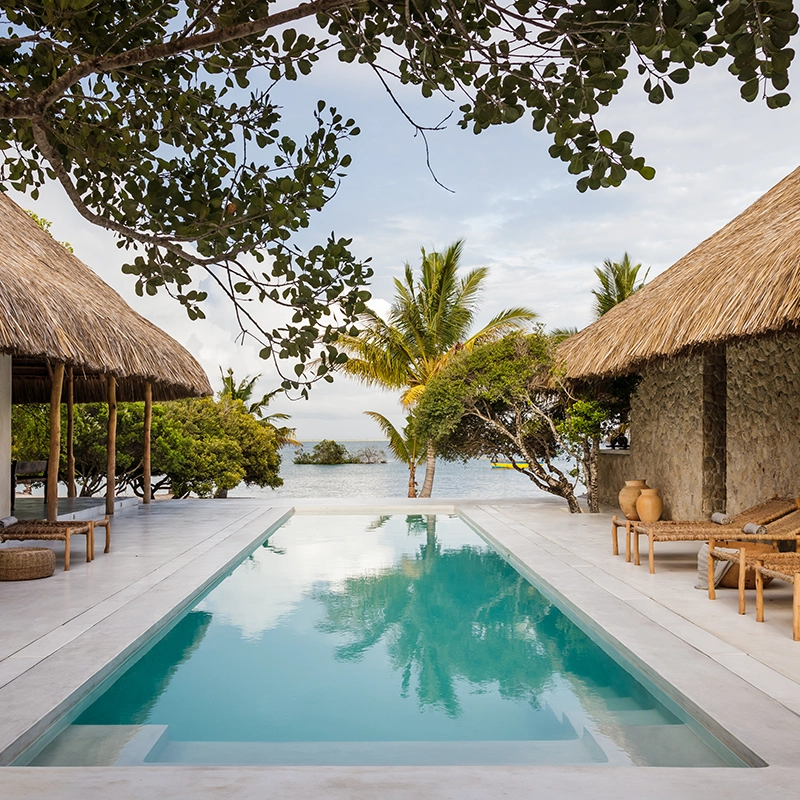 Perfect Hideaways: A serene beachfront pool framed by thatched-roof villas, woven loungers and lush palms, opening up to tranquil blue waters under a soft afternoon sky. Sussurro, Vilanculos.