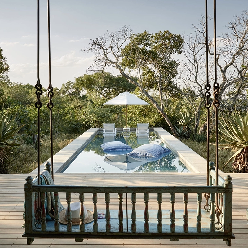 Perfect Hideaways – A serene outdoor pool deck set against tall golden grasses and a soft sunset glow. Two bright red chairs face the still, reflective water, with a small clear side table between them holding two glasses of champagne. To the left, two cushioned wooden loungers with rolled white towels sit beneath a shaded structure. Beyond the deck, lush greenery stretches toward the horizon as warm sunlight filters through the trees. Royal Malewane Farmstead, Thornybush Game Reserve