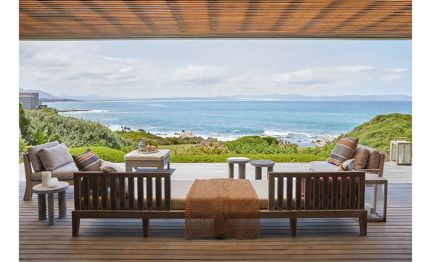Perfect Hideaways: A tranquil outdoor lounge with timber seating and neutral-toned cushions looks out across a lush garden to a sweeping view of the turquoise sea, framed by a timber-slatted pergola. Cliffside, Hermanus.