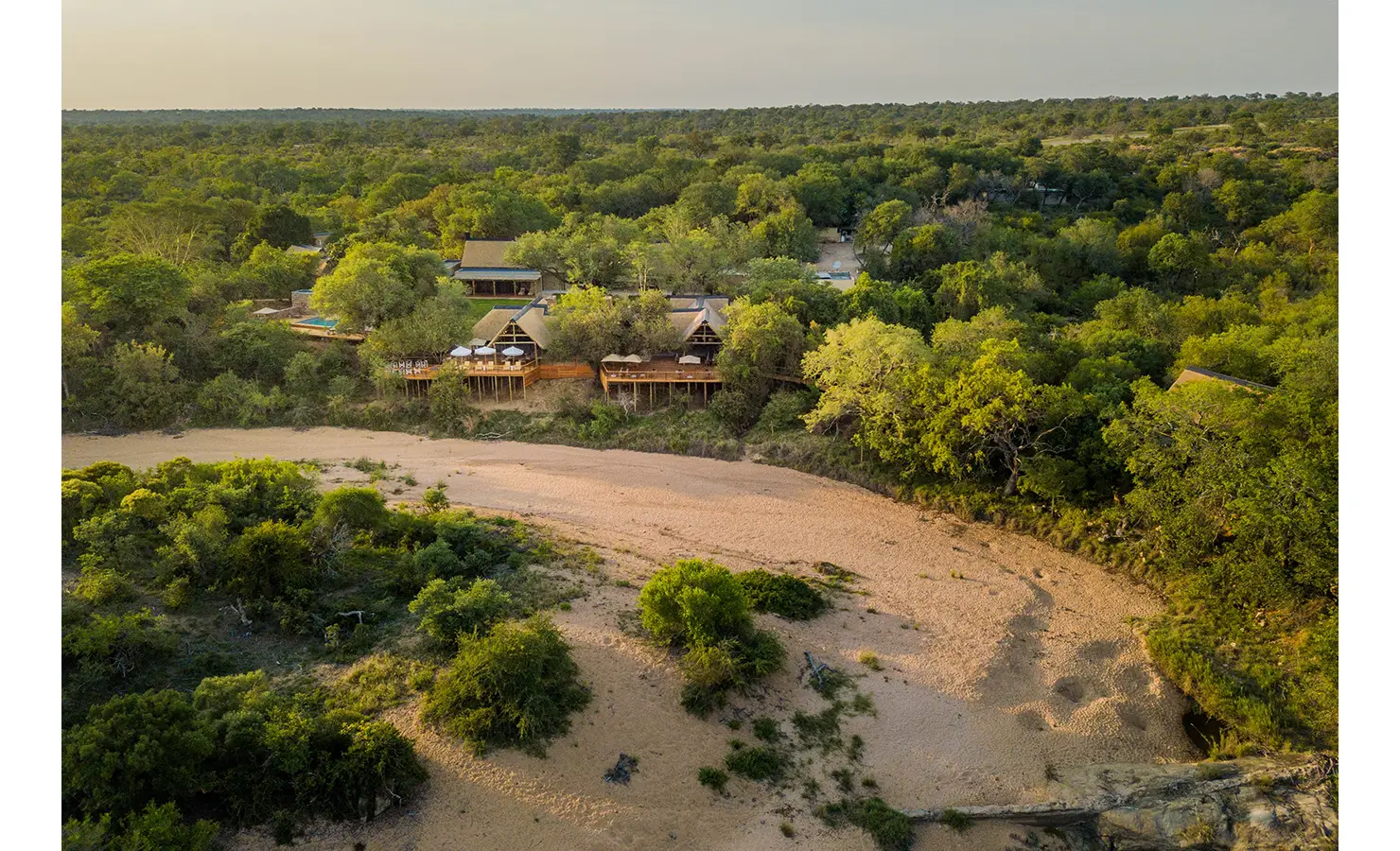 Perfect Hideaways: An elevated view of Thornybush Lodge, surrounded by dense bushveld with thatched-roof chalets and wooden decks overlooking a dry riverbed. The lodge’s open-plan design blends seamlessly with the wilderness, creating a serene and immersive safari retreat. Thornybush Lodge, Thornybush Game Reserve.