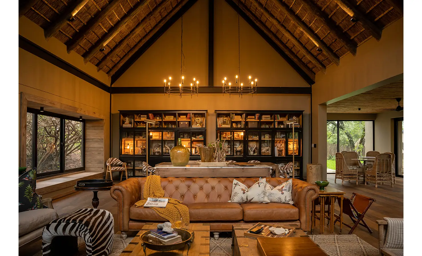 Perfect Hideaways: A stylish library and dining space at Thornybush Lodge, featuring a floor-to-ceiling display of books and curiosities. A guest browses the collection, while a rustic wooden dining table with zebra-print chairs adds a touch of safari elegance. Thornybush Lodge, Thornybush Game Reserve.