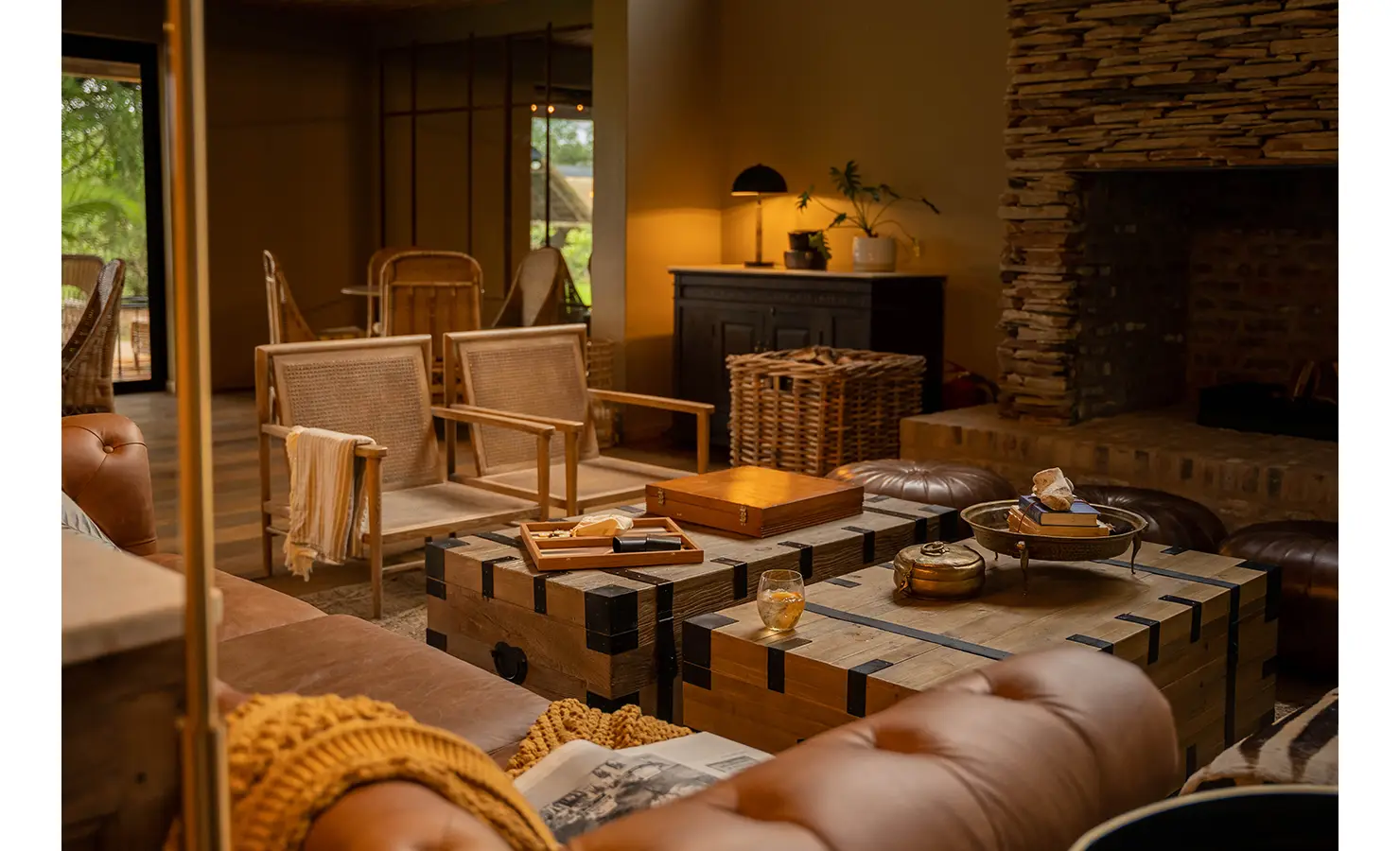 Perfect Hideaways: A cosy lounge area illuminated by warm chandeliers, with plush leather seating and a wall of glass-fronted cabinets showcasing books and artefacts. Large windows frame views of the surrounding wilderness. Thornybush Lodge, Thornybush Game Reserve.