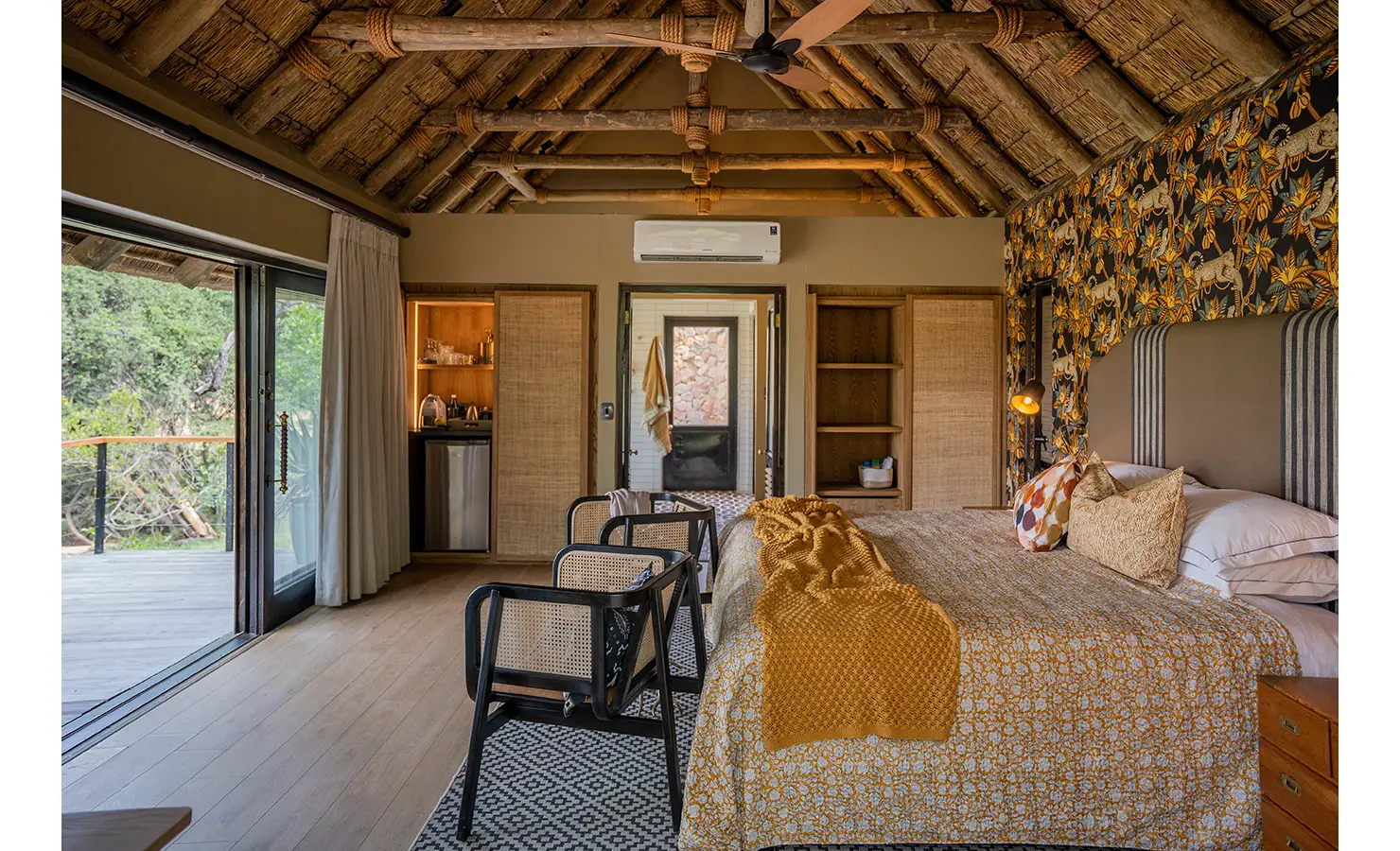 Perfect Hideaways: A beautifully designed bedroom with a thatched ceiling, featuring bold wildlife-inspired wallpaper, a plush bed with warm-toned textiles, and an open doorway leading to the surrounding bush. Thornybush Lodge, Thornybush Game Reserve.