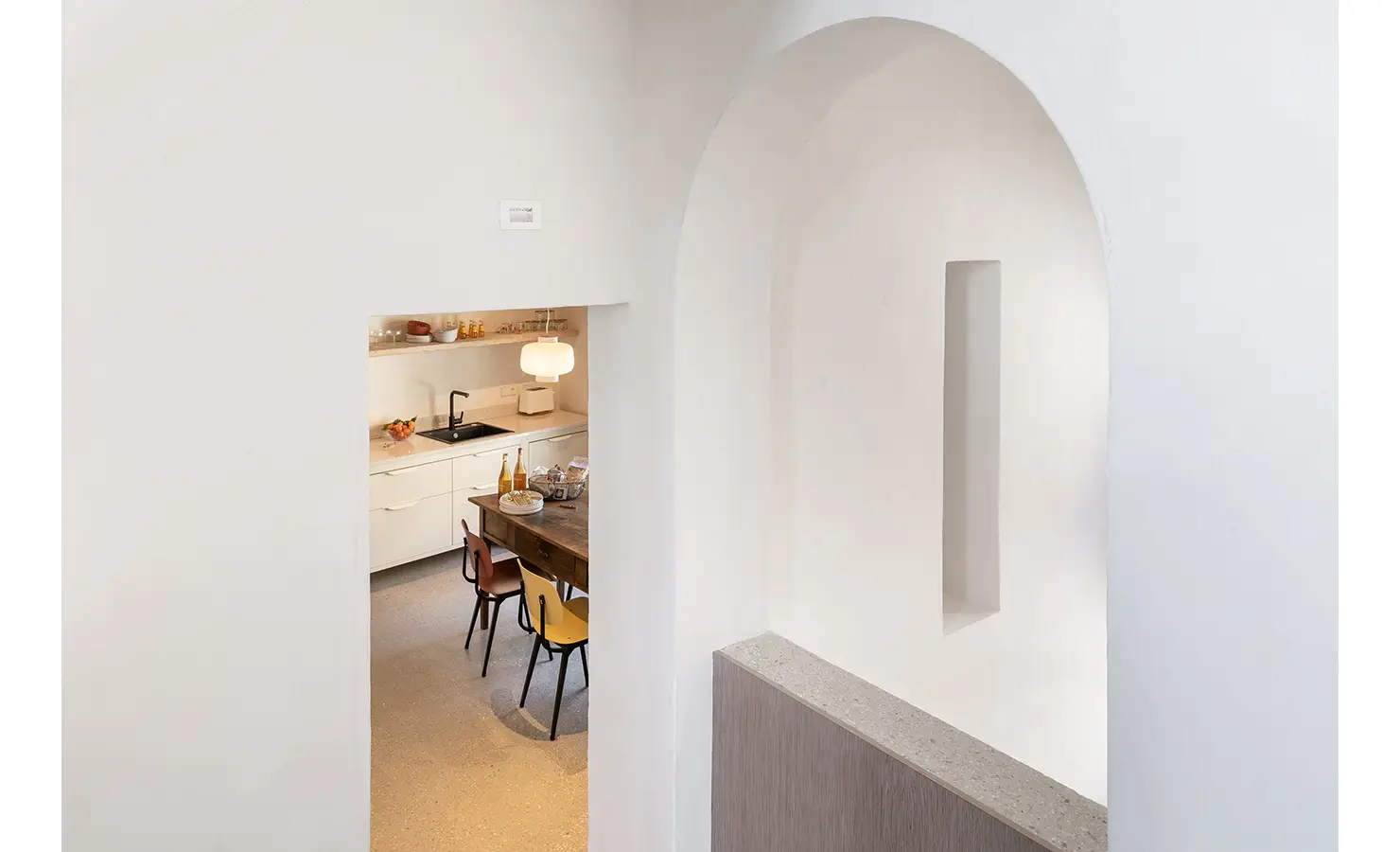 Perfect Hideaways: A view of the kitchen from a white arched hallway, showcasing the open-plan design with natural light streaming through large windows. Umbria Cottage, Collazzone.