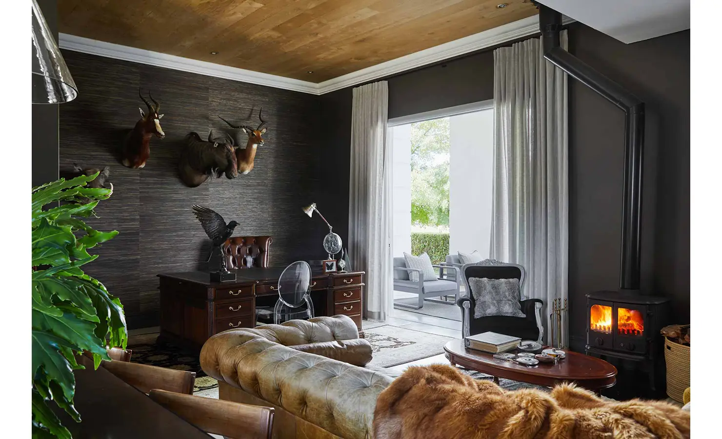 Perfect Hideaways: A richly decorated study and lounge area with a leather Chesterfield, mounted antelope heads, a traditional wooden desk, and a cosy wood-burning stove beneath a timber ceiling. Brenaissance Winelands Manor House, Stellenbosch.