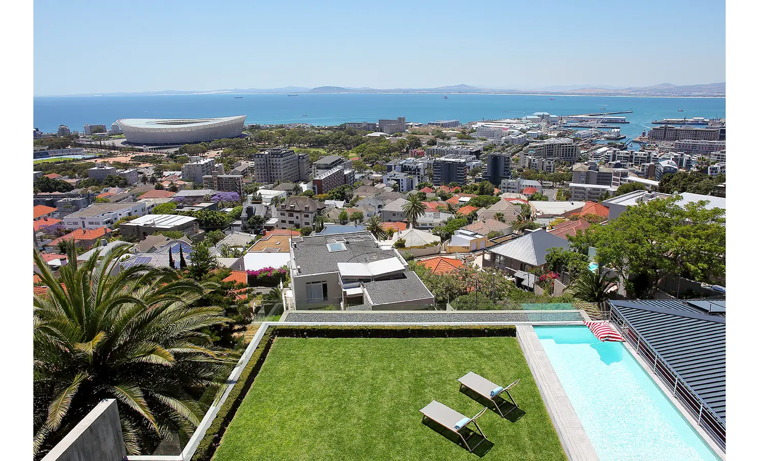 Perfect Hideaways: A lush green lawn and elevated swimming pool overlook the city’s rooftops and coastline, with the Cape Town Stadium and ocean forming a dramatic backdrop. Hawk's Eye, Green Point.