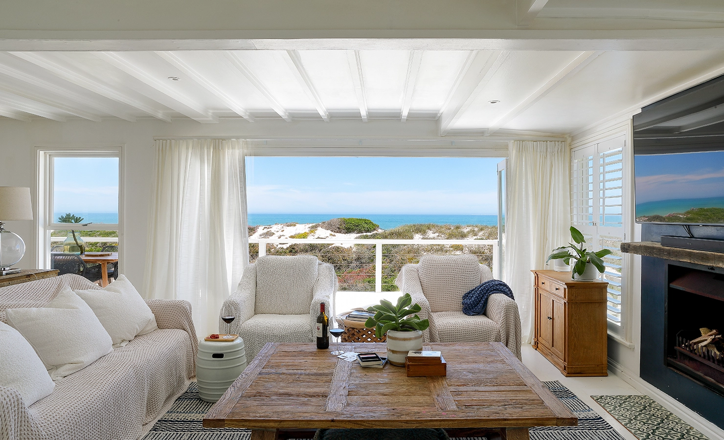 Light and spacious lounge that reflects in the furniture of Sandpiper with soft, beige sofas and arm chairs that surround a large wooden coffee table and facing a built-in fire place opening onto views of the coastal dunes and ocean from large stacking doors.