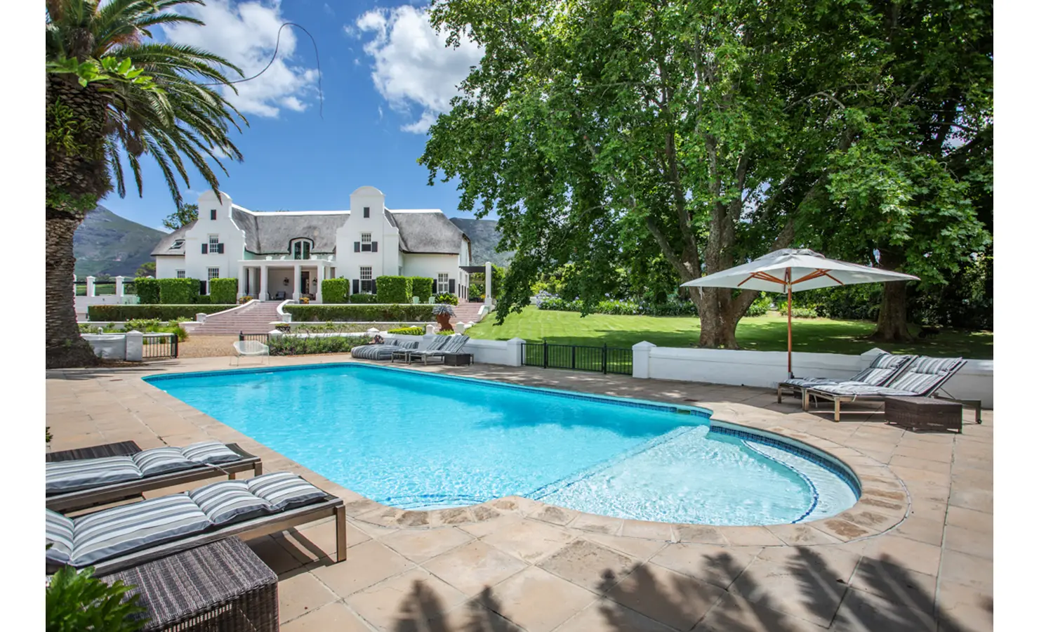 05| Perfect Hideaways: A large swimming pool set in a sun-soaked patio is framed by palm trees, striped loungers, and expansive lawns leading up to a gabled Cape Dutch house. Buitenzorg Pool Cottage, Constantia.
