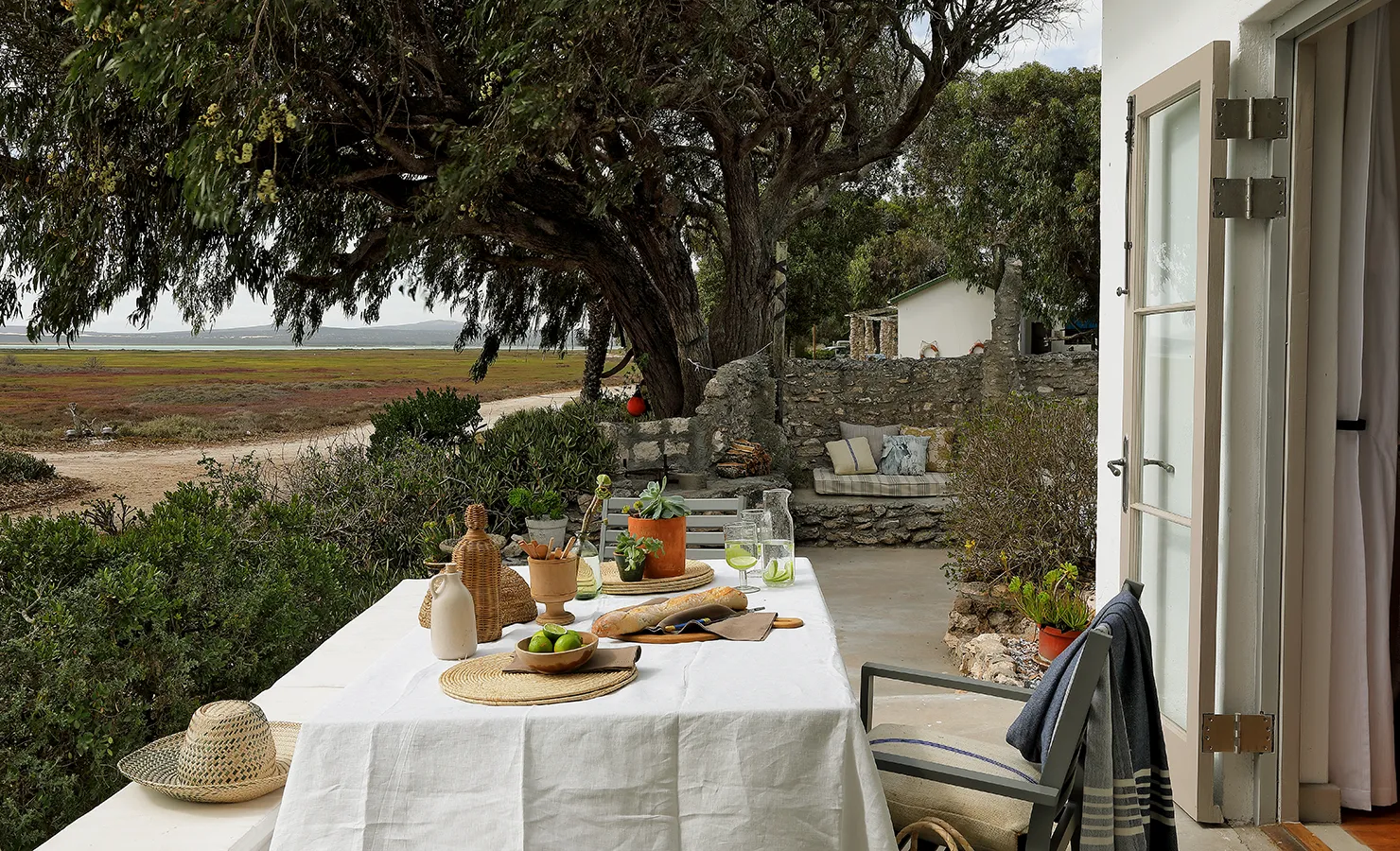 Perfect Hideaways: A rustic outdoor dining table set with fresh produce and natural decor, positioned under the shade of a large tree with views of the surrounding landscape. Berit's Cottage, Churchhaven.