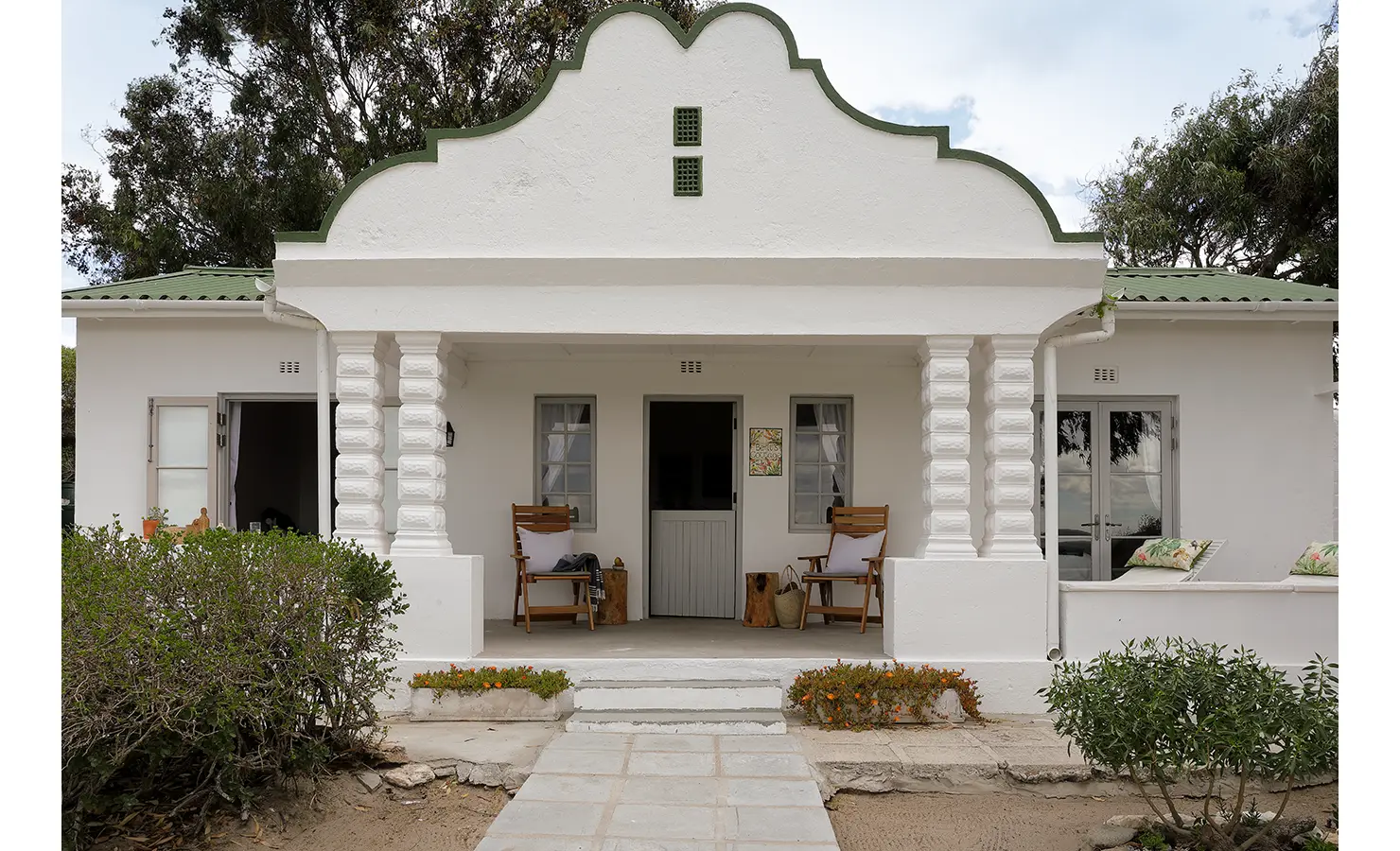 Perfect Hideaways: A traditional whitewashed cottage facade with curved gable detailing, flanked by lush greenery and featuring a welcoming entrance. Berit's Cottage, Churchhaven.