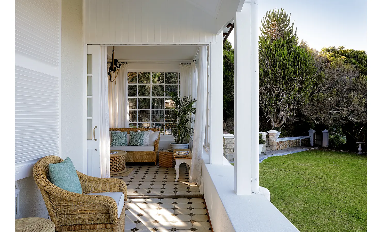 Perfect Hideaways: A charming covered veranda with wicker armchairs and a tiled floor, opening onto a lush green lawn with a backdrop of native trees and foliage. Flowing white curtains frame a sunlit seating area, creating an inviting indoor-outdoor retreat. Boulders House, Simons Town.