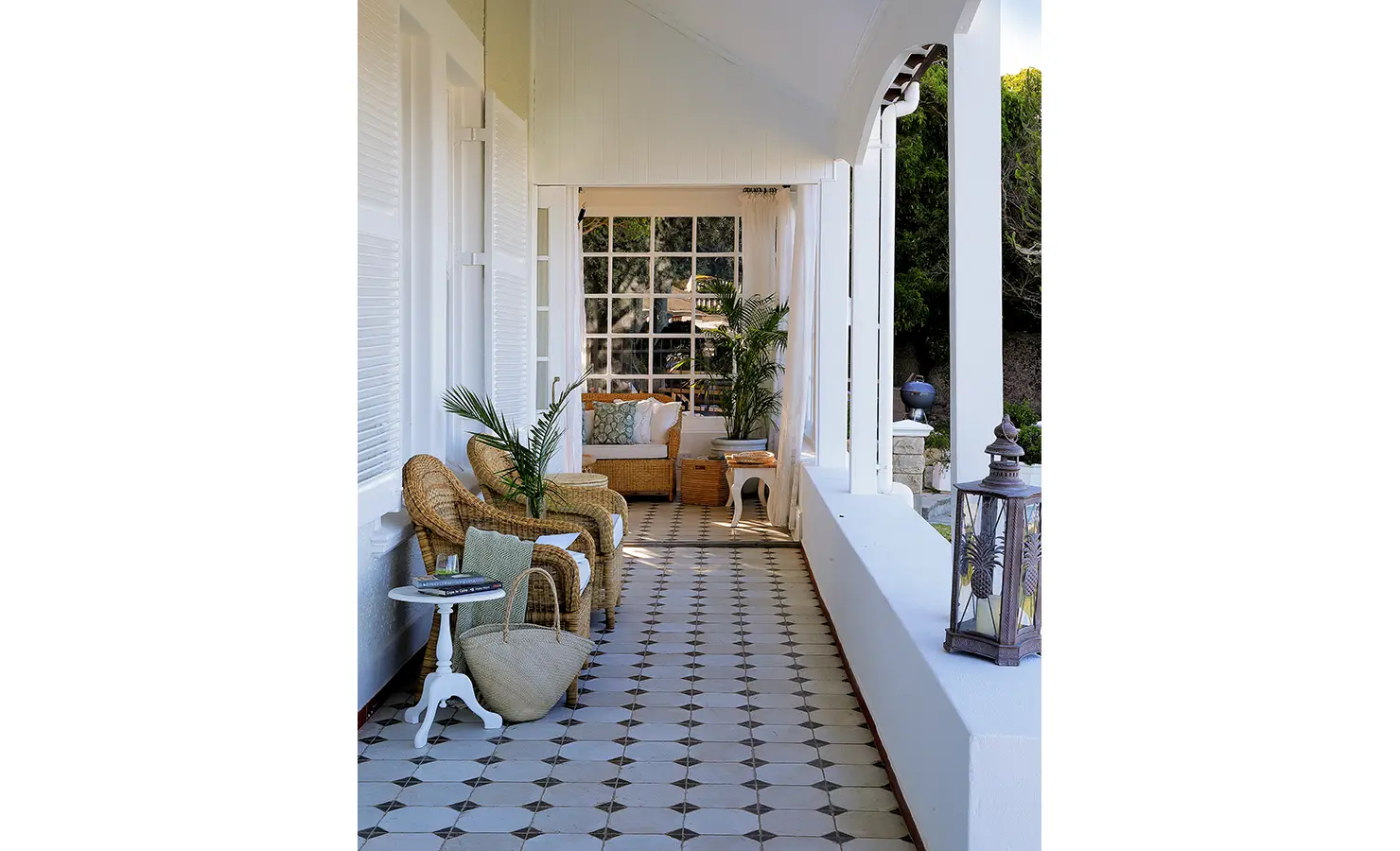 Perfect Hideaways: A beautifully styled veranda with a row of wicker chairs, soft cushions, and potted greenery, leading to a cosy lounge framed by sheer white curtains. The elegant black-and-white tiled floor enhances the classic coastal charm. Boulders House, Simons Town.