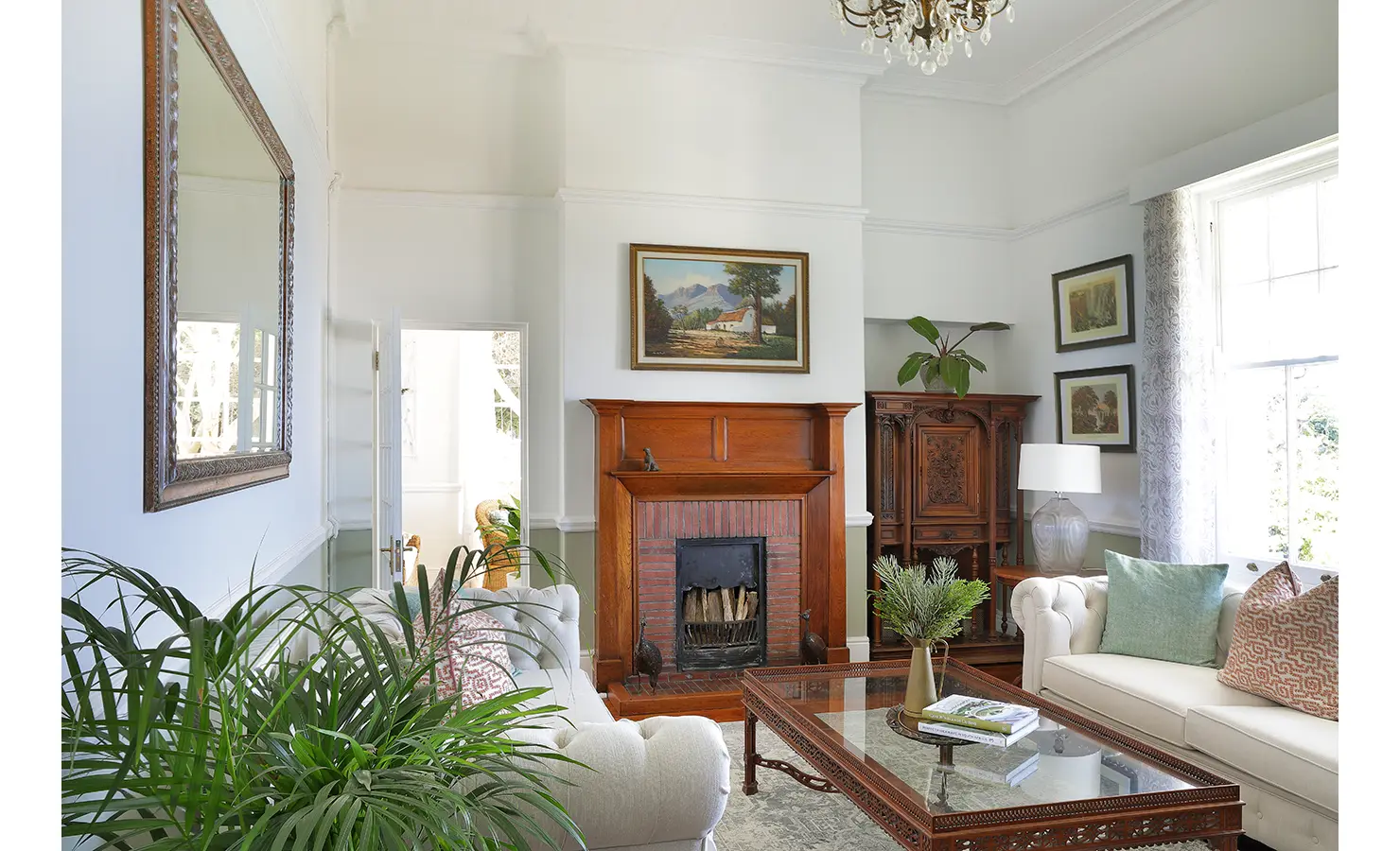 Perfect Hideaways: A refined living room blending classic and contemporary elements, featuring a rich wooden fireplace, an antique cabinet, and plush cream sofas with pastel accents. Ornate framed artwork and a sparkling chandelier complete the timeless décor. Boulders House, Simons Town.