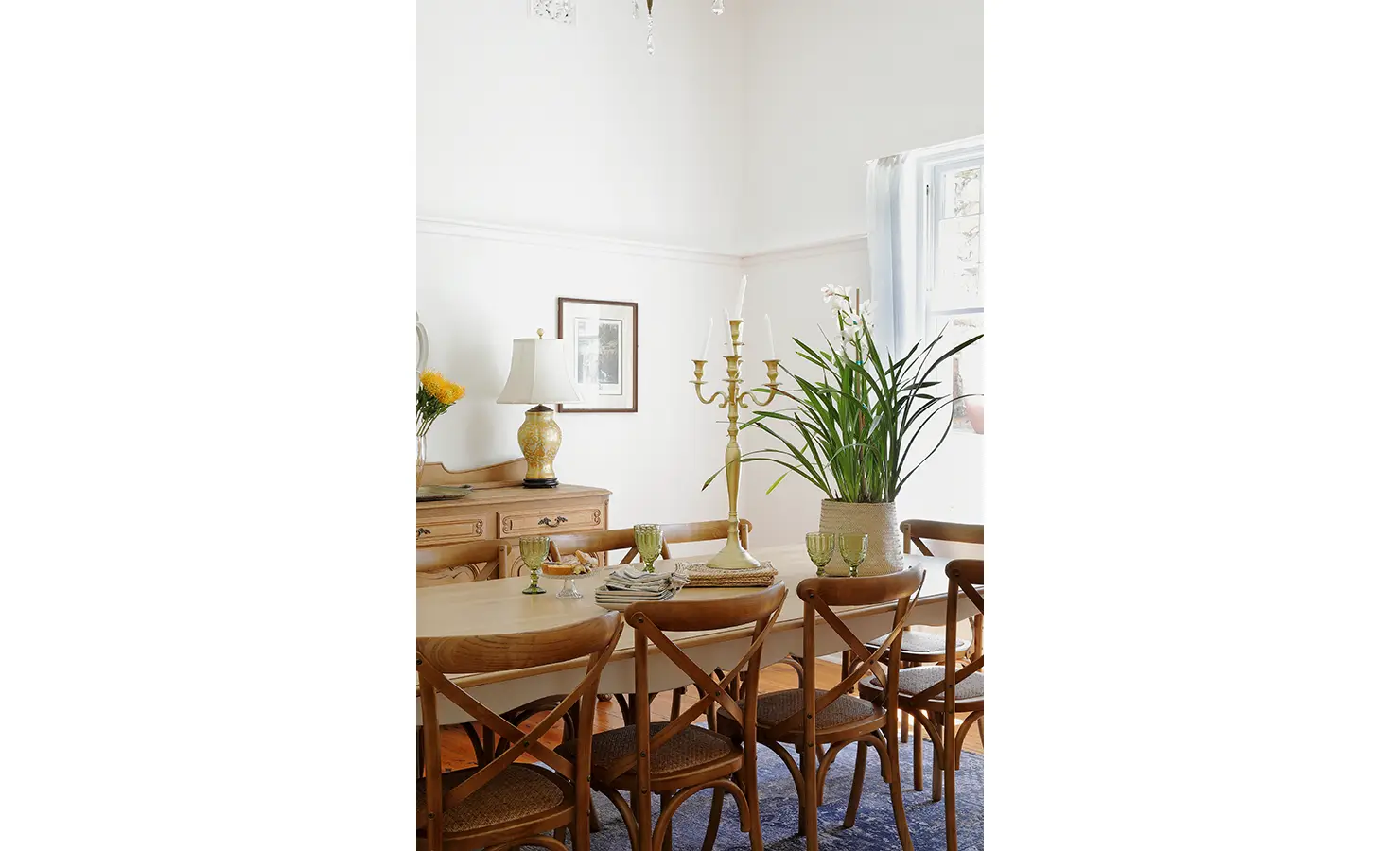 Perfect Hideaways: A bright and airy dining room with a long wooden table surrounded by classic cross-back chairs. A soft colour palette, fresh greenery, and a vintage candelabra create a warm, inviting atmosphere. Boulders House, Simons Town.