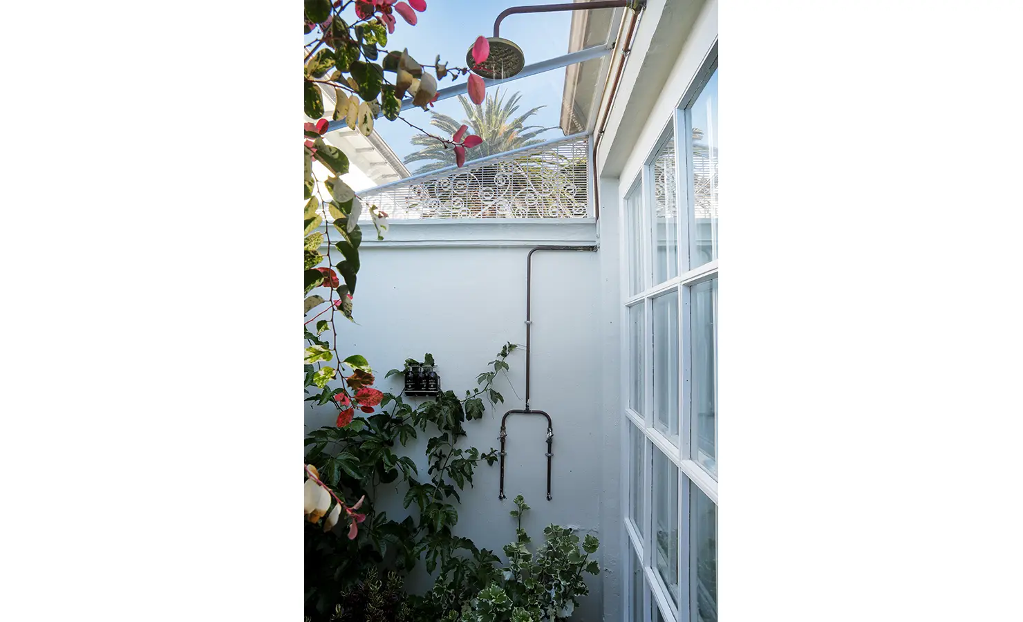 Perfect Hideaways: A secluded outdoor shower enclosed by leafy greenery, featuring a rustic black metal showerhead. The delicate white trellis detail adds a touch of elegance to this peaceful alfresco space. Boulders House, Simons Town.