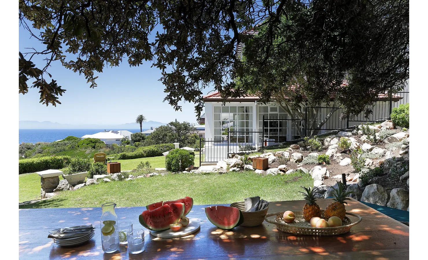 Perfect Hideaways: A sun-dappled outdoor dining spot beneath a shady tree, with a spread of fresh watermelon, pineapples, and citrus fruits. The lush garden and distant ocean views enhance the relaxed coastal ambiance. Boulders House, Simons Town.