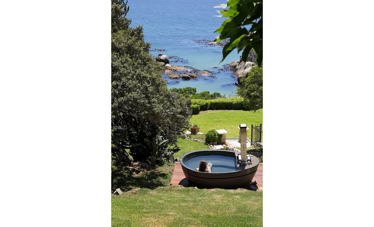 Perfect Hideaways: A wood-fired hot tub set in a terraced garden, offering a panoramic view of the deep blue sea and lush coastal greenery. A perfect spot for relaxation with nature. Boulders House, Simons Town.