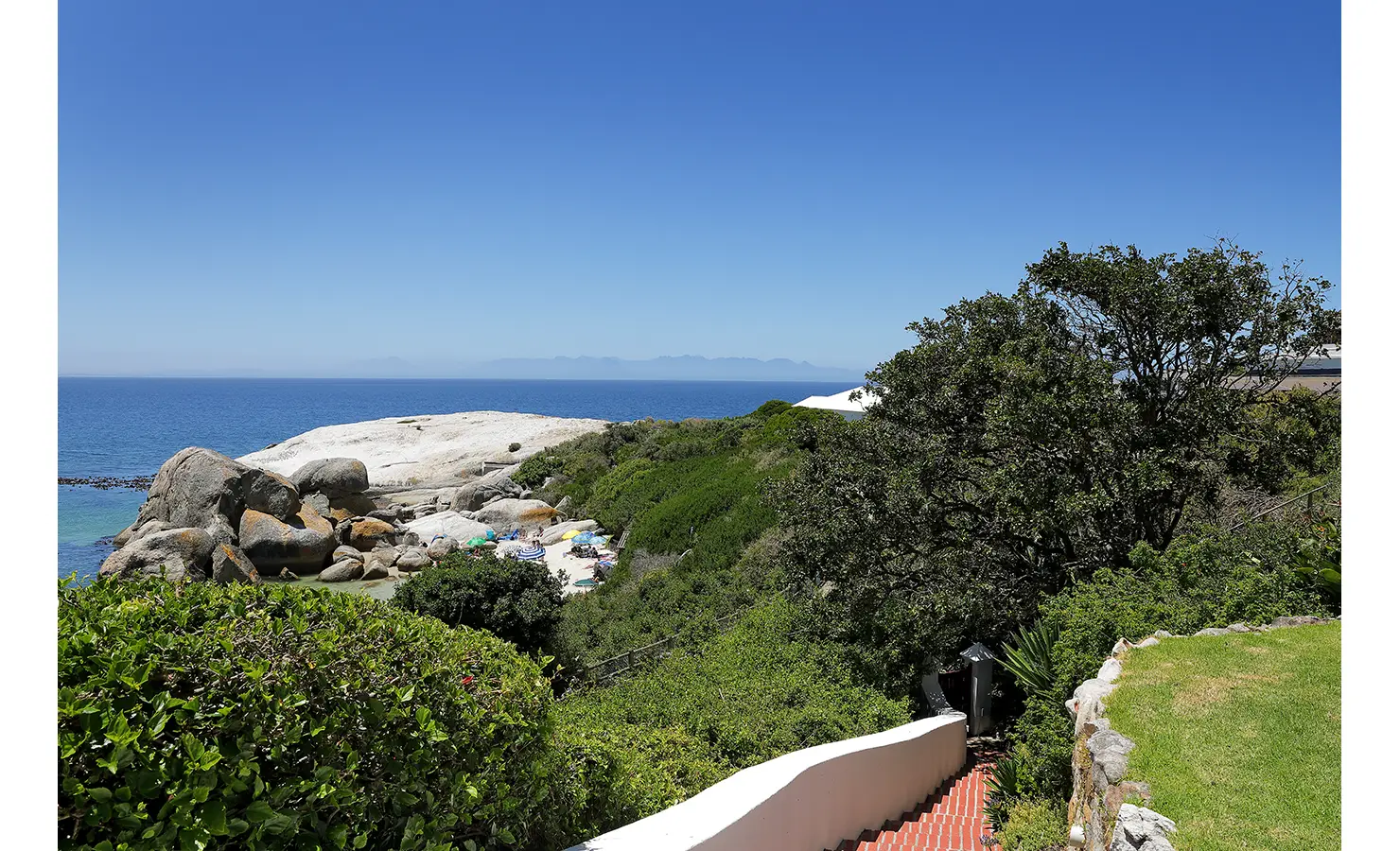Perfect Hideaways: A scenic pathway with terracotta steps winding through the greenery, leading to a secluded beach framed by smooth granite boulders and the sparkling ocean. Boulders House, Simons Town.
