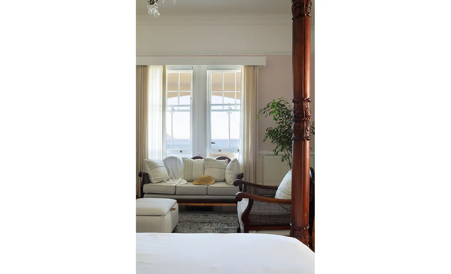 Perfect Hideaways: A sunlit bedroom corner with a plush cream sofa and ocean views framed by tall sash windows. Soft drapes and warm textiles invite relaxation in this tranquil coastal sanctuary. Boulders House, Simons Town.
