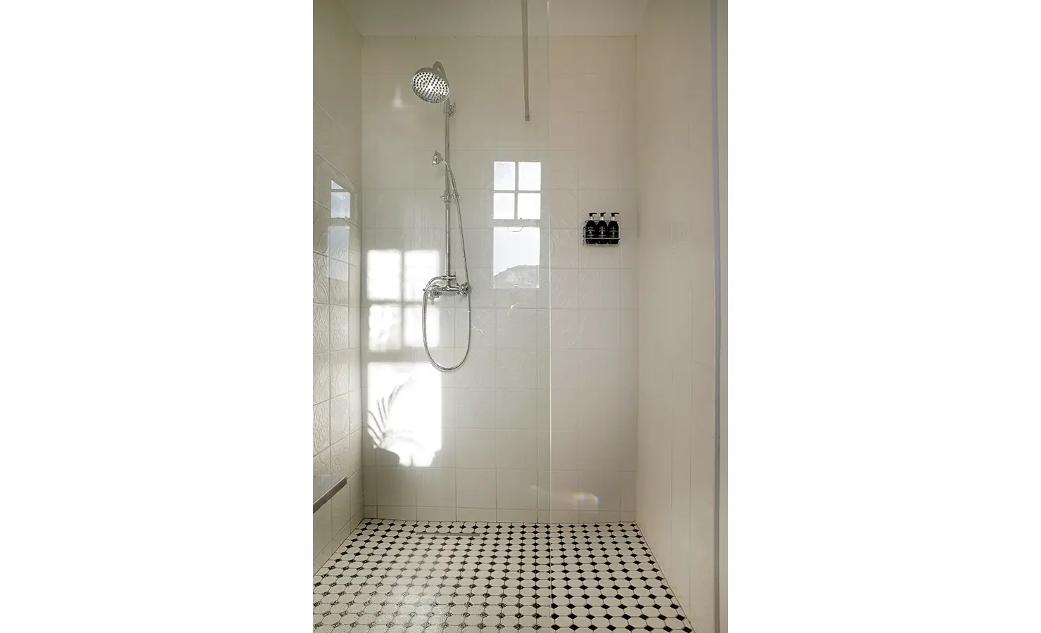 Perfect Hideaways: A minimalist white-tiled shower with vintage-inspired black-and-white floor tiles, bathed in soft natural light from a nearby window. A modern yet timeless space for relaxation. Boulders House, Simons Town.
