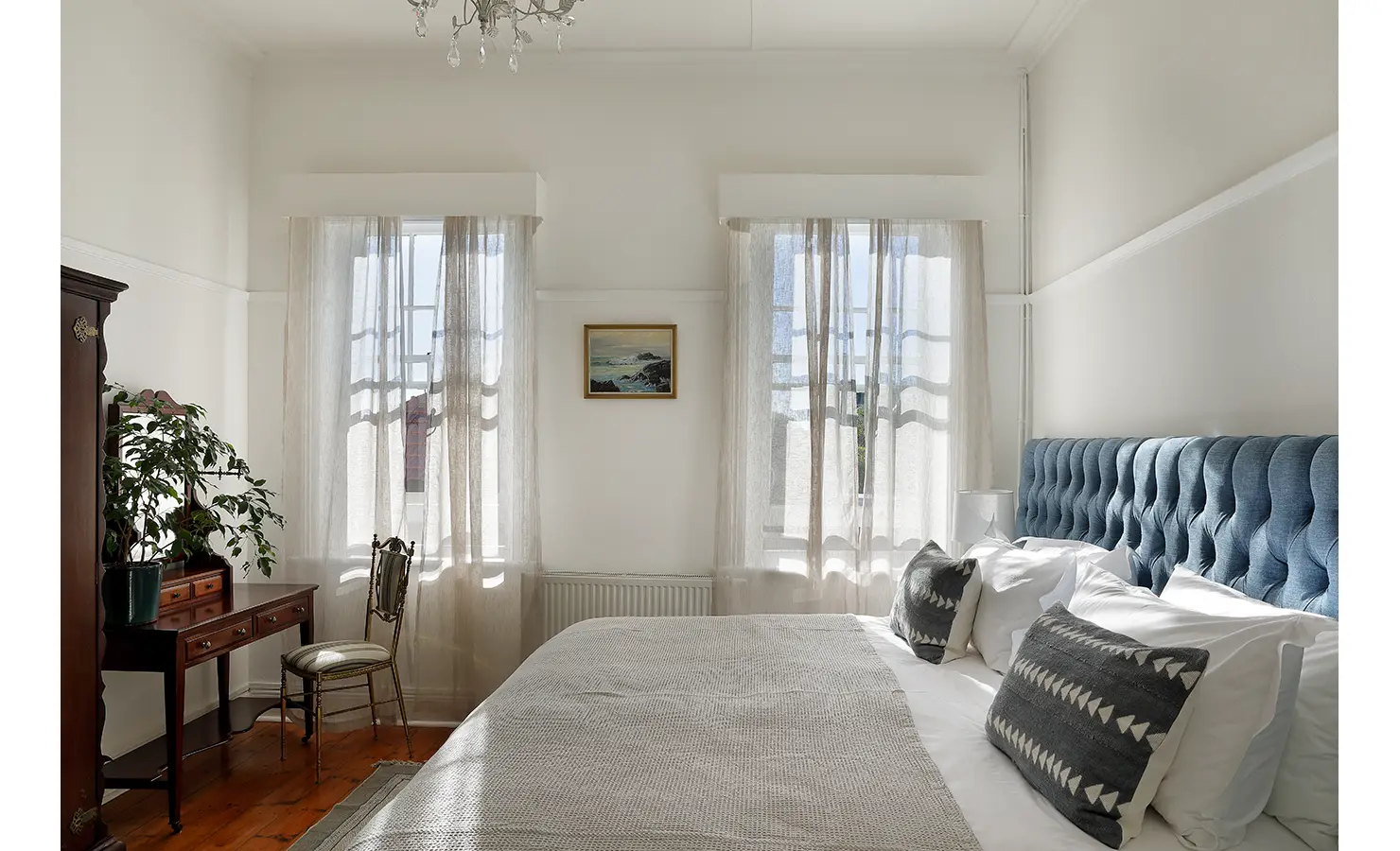 Perfect Hideaways: A bright bedroom with a deep blue upholstered headboard, antique furnishings, and sheer white curtains allowing sunlight to filter through. The peaceful space is softened with neutral tones and natural textures. Boulders House, Simons Town.