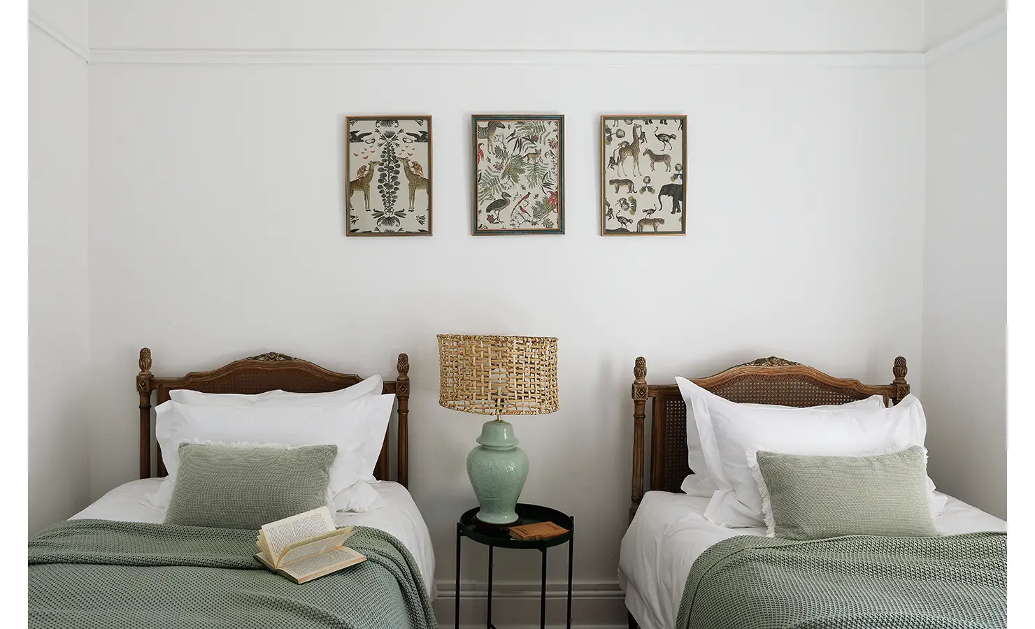 Perfect Hideaways: A twin bedroom with elegant wooden headboards, soft green blankets, and framed nature-inspired artwork above the beds. A woven lamp adds a touch of rustic charm. Boulders House, Simons Town.