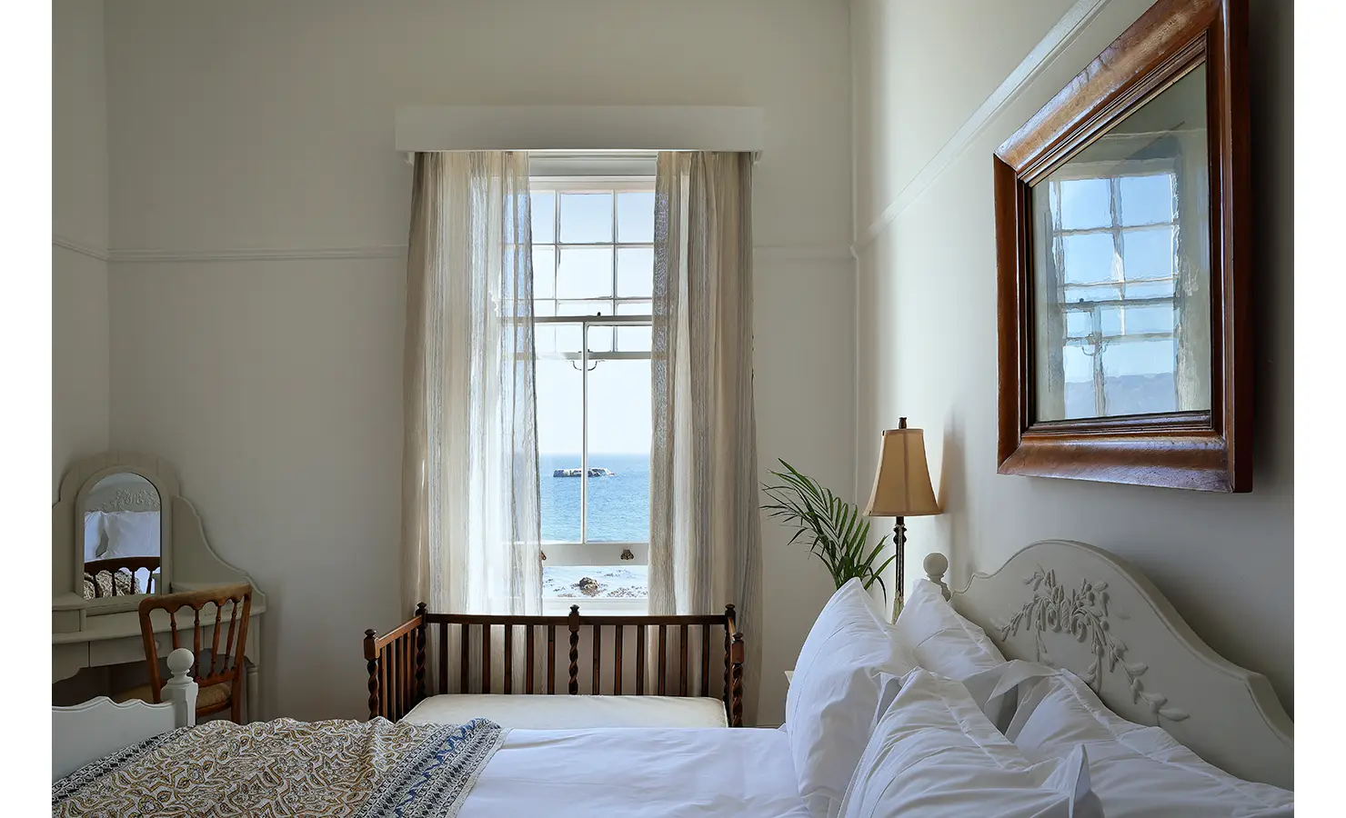 Perfect Hideaways: A dreamy bedroom with a white vintage-style bed, a delicate writing desk, and a sash window framing breathtaking ocean views. Sheer curtains and neutral tones add to the tranquil atmosphere. Boulders House, Simons Town.