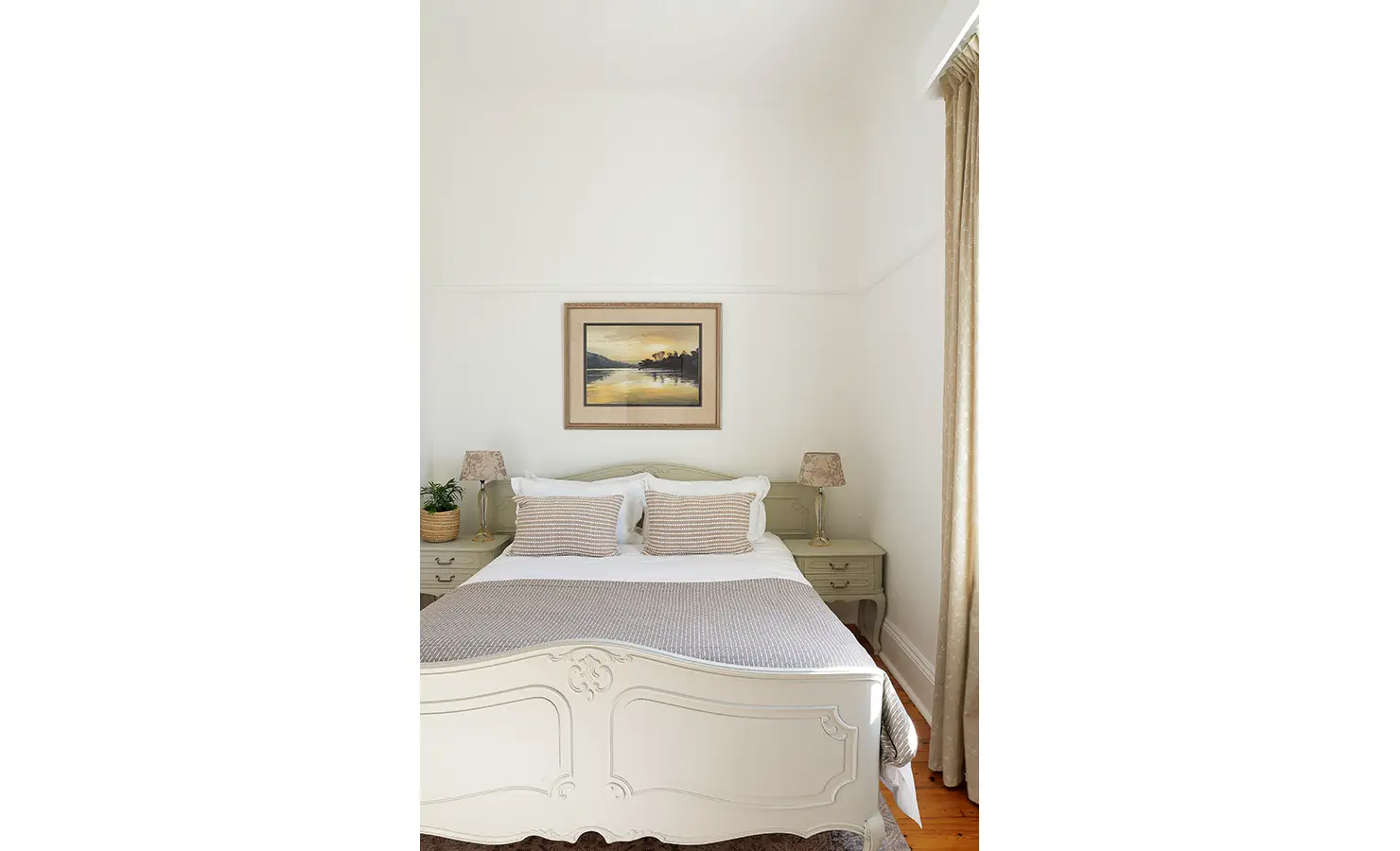 Perfect Hideaways: A charming bedroom with a soft cream bed frame, warm neutral bedding, and antique-style bedside tables. A landscape painting above the bed completes the inviting aesthetic. Boulders House, Simons Town.