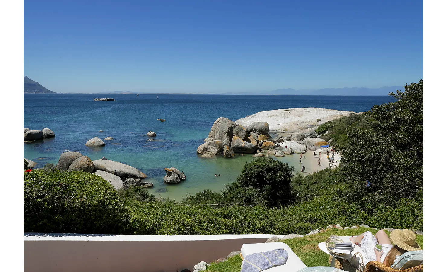 Perfect Hideaways: A sun-drenched terrace with an uninterrupted view of the bay, where the azure waters meet the horizon. A wicker lounger with soft cushions offers the perfect spot for seaside relaxation. Boulders House, Simons Town.