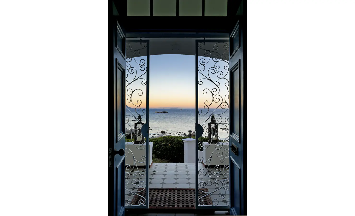 Perfect Hideaways: A breathtaking view from the entrance, framed by ornate wrought iron gates, looking out to the shimmering ocean at sunset. A moment of pure tranquillity. Boulders House, Simons Town.