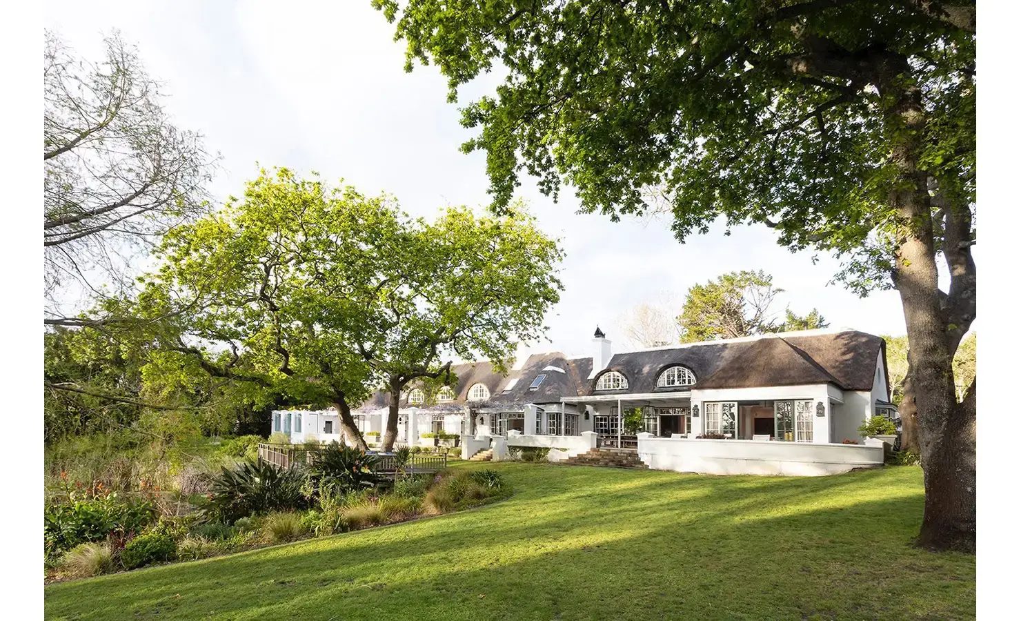 Front view of The Old Manor House with thatch roof, expansive garden and large trees.
