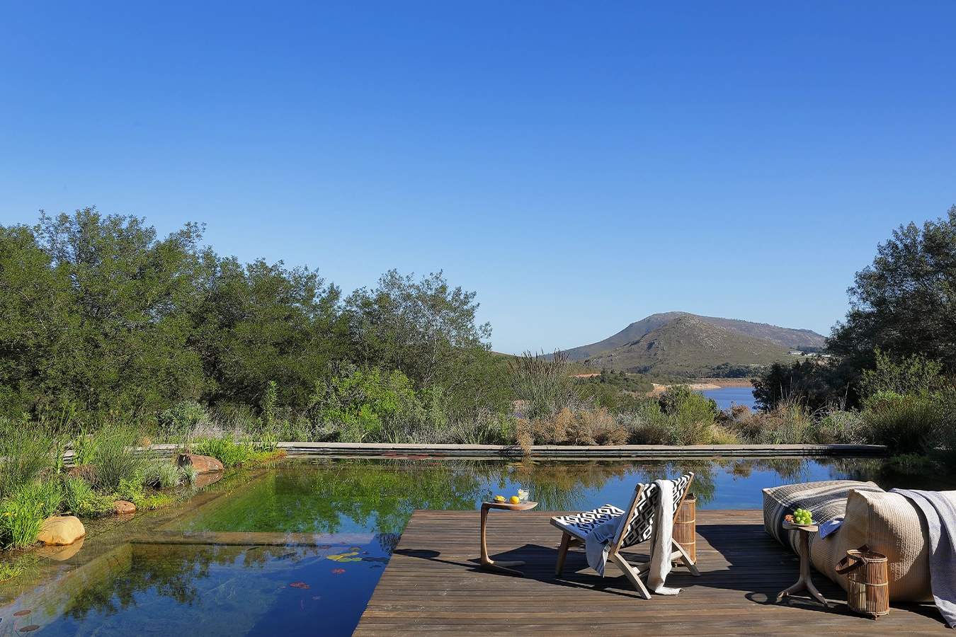 Perfect Hideaways: A serene natural swimming pool with wooden deck, sun loungers and mountain views across the Hemel-en-Aarde Valley, surrounded by lush greenery and open skies. Stonehaven House, Hemel-en-Aarde.