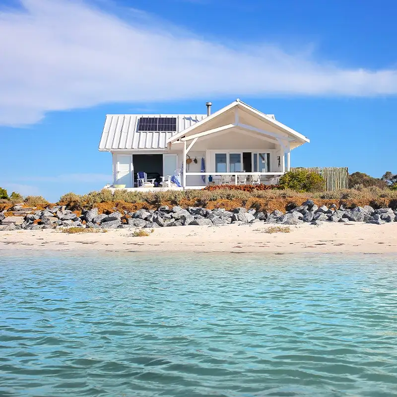 Perfect Hideaways: A cozy white beach house with solar panels sits by a rocky shoreline, under a clear blue sky. Calm turquoise waters create a serene atmosphere. Shoot Locations.