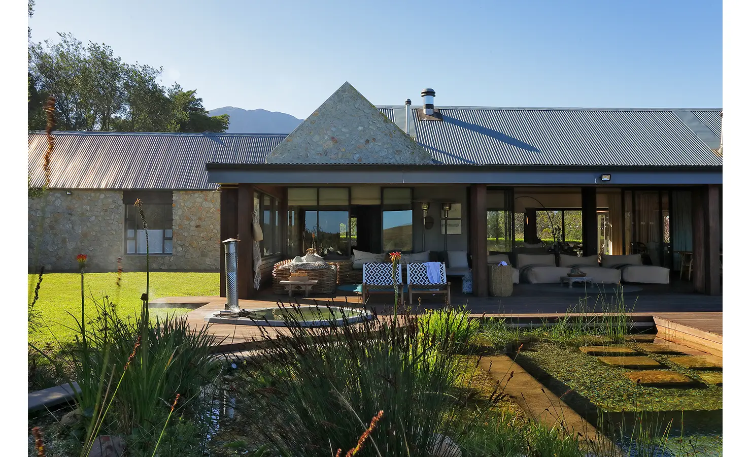 Perfect Hideaways: Stone-clad exterior with shaded veranda and cosy outdoor seating overlooking a natural pond and mountain backdrop. Stonehaven House, Hemel-en-Aarde.