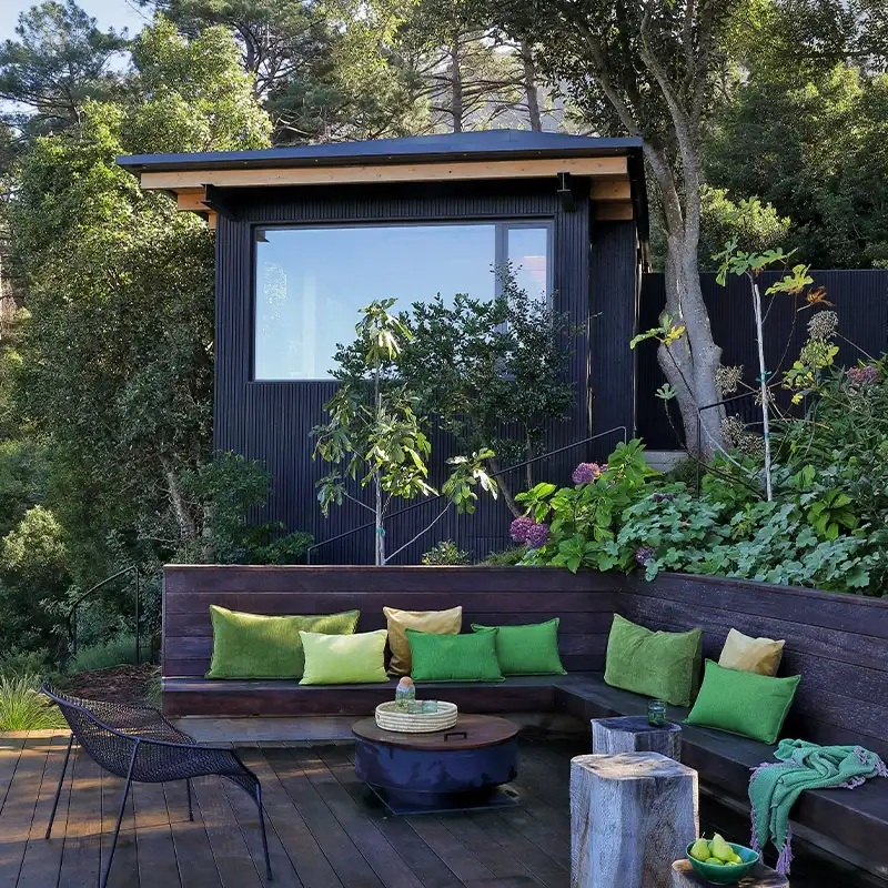 Perfect Hideaways: Secluded outdoor deck with cozy seating area featuring green and yellow cushions, surrounded by lush foliage and trees, creating a serene ambiance. Shoot Locations.