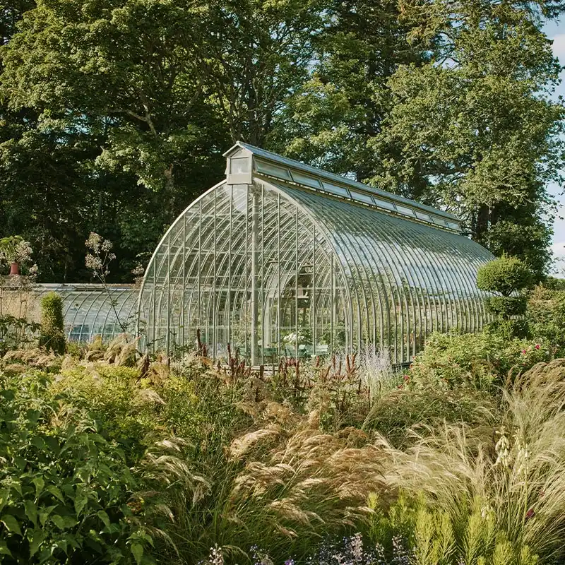Perfect Hideaways: A glass greenhouse stands amidst a lush garden with various vibrant plants and grasses. Tall trees surround the scene under a clear, blue sky. Shoot Locations.