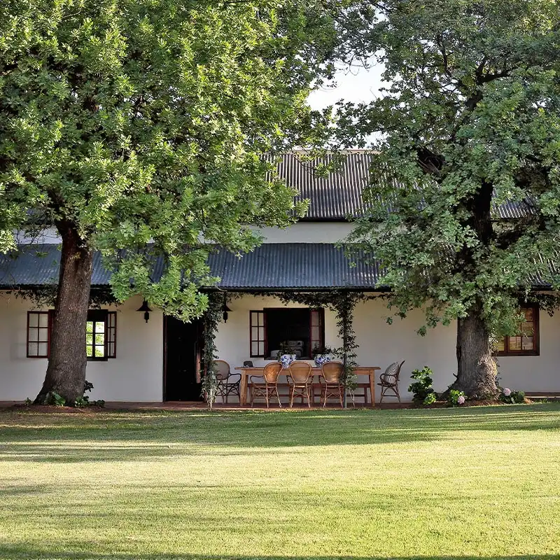 Perfect Hideaways: A charming country house with a corrugated metal roof, flanked by two large trees. A wooden table and chairs sit on the grass, evoking a serene, rustic feel. Shoot Locations.