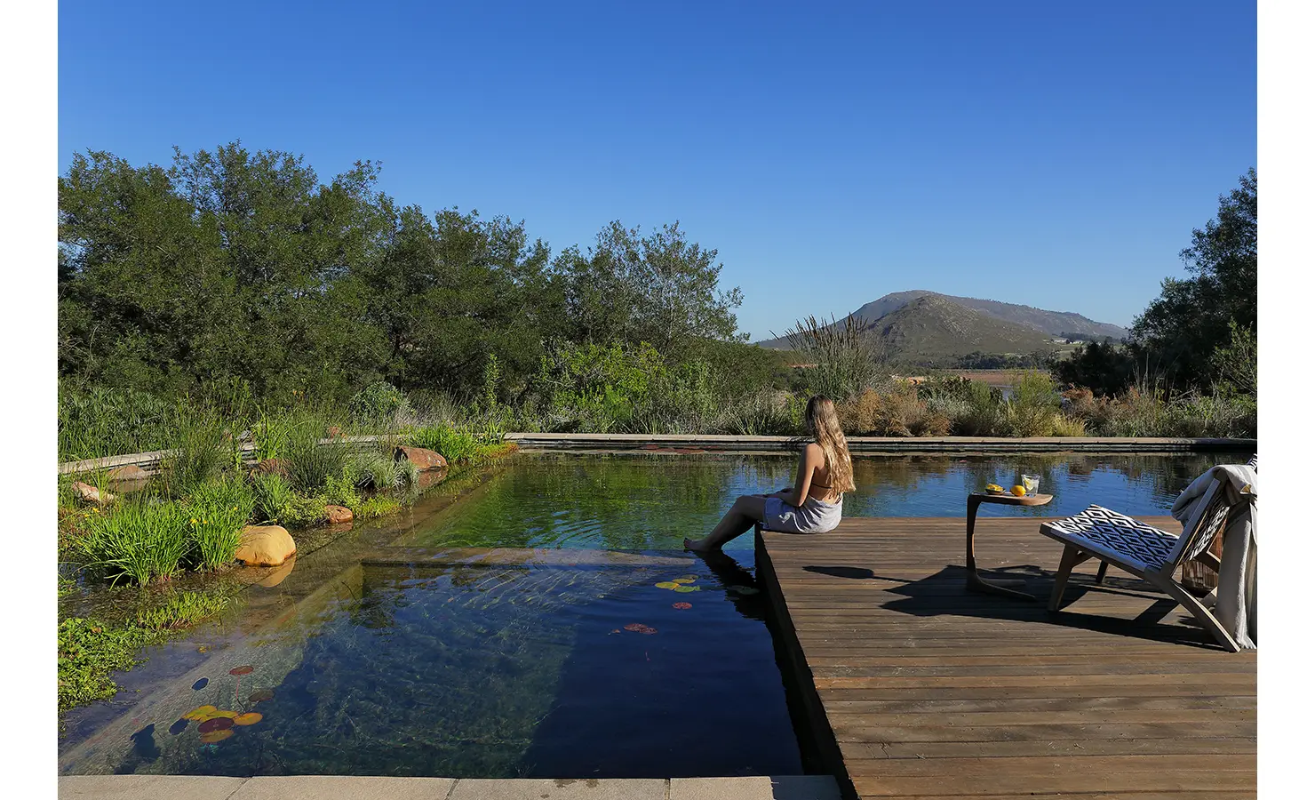 Perfect Hideaways: Relaxed moment by the natural pool as a guest dangles her feet in the water with mountain scenery beyond. Stonehaven House, Hemel-en-Aarde.