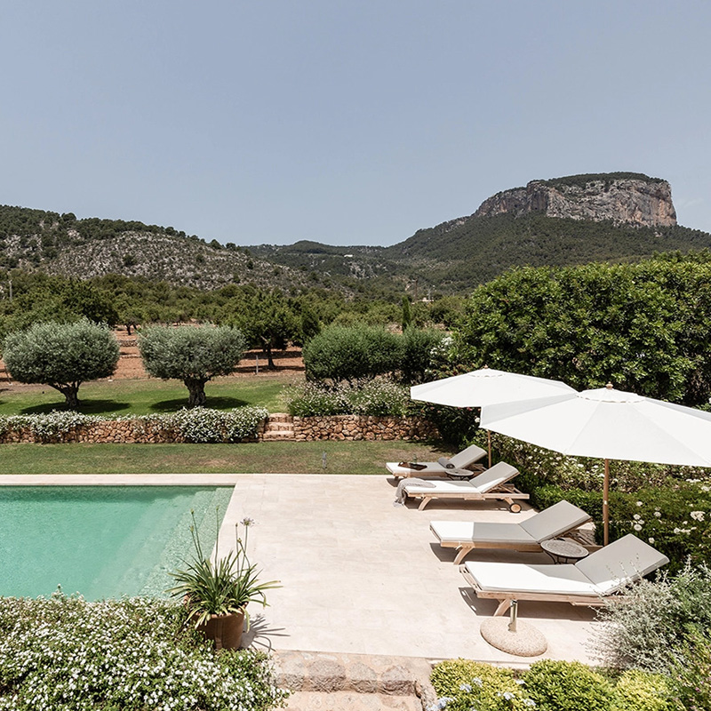 Perfect Hideaways – A tranquil pool terrace overlooking manicured lawns, olive trees, and rolling Mallorcan mountains beneath a clear sky, with sun loungers and umbrellas arranged along the stone poolside – Finca Es Rafal, Alaró