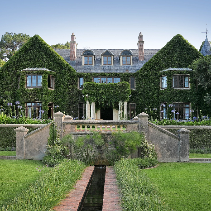 Perfect Hideaways — A grand ivy-covered manor house with elegant symmetrical architecture, tall chimneys, and classic wooden windows surrounded by lush greenery. The home features a formal garden with manicured lawns, flowering agapanthus, and a reflective water feature leading toward the stately entrance with columns and a stone balustrade. A serene, luxurious retreat set against mature trees under a clear blue sky. Bishopscourt, Bishops Hall.