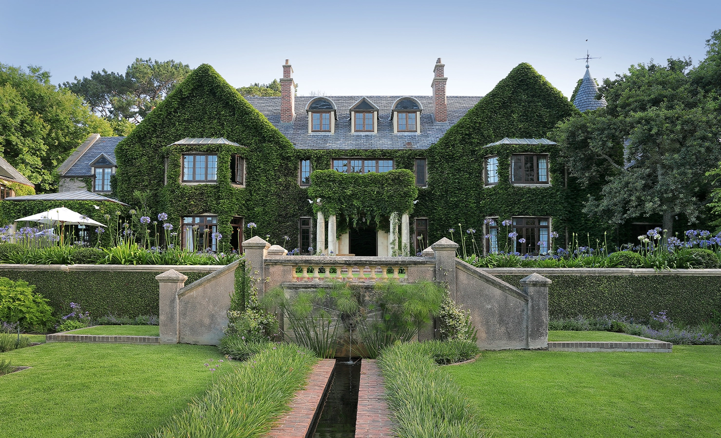 Perfect Hideaways — A grand ivy-covered manor house with elegant symmetrical architecture, tall chimneys, and classic wooden windows surrounded by lush greenery. The home features a formal garden with manicured lawns, flowering agapanthus, and a reflective water feature leading toward the stately entrance with columns and a stone balustrade. A serene, luxurious retreat set against mature trees under a clear blue sky. Bishopscourt, Bishops Hall.