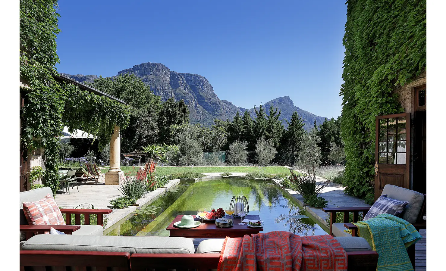 Perfect Hideaways — A tranquil courtyard with a natural swimming pool surrounded by lush greenery and mountain views. Comfortable outdoor seating and a table set for breakfast create a relaxed, elegant ambiance under a vivid blue sky. Bishopscourt, Bishops Hall.
