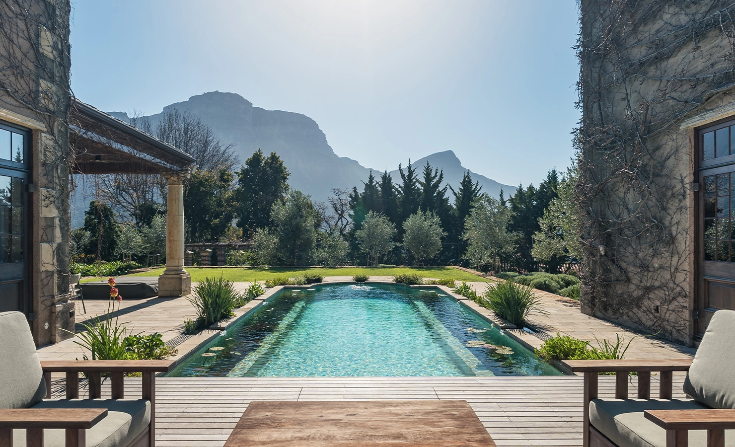 Perfect Hideaways: Tranquil swimming pool framed by stone buildings and wooden loungers, leading toward manicured lawns, tall trees and dramatic mountain views under clear blue skies, Bishopscourt, Bishops Hall