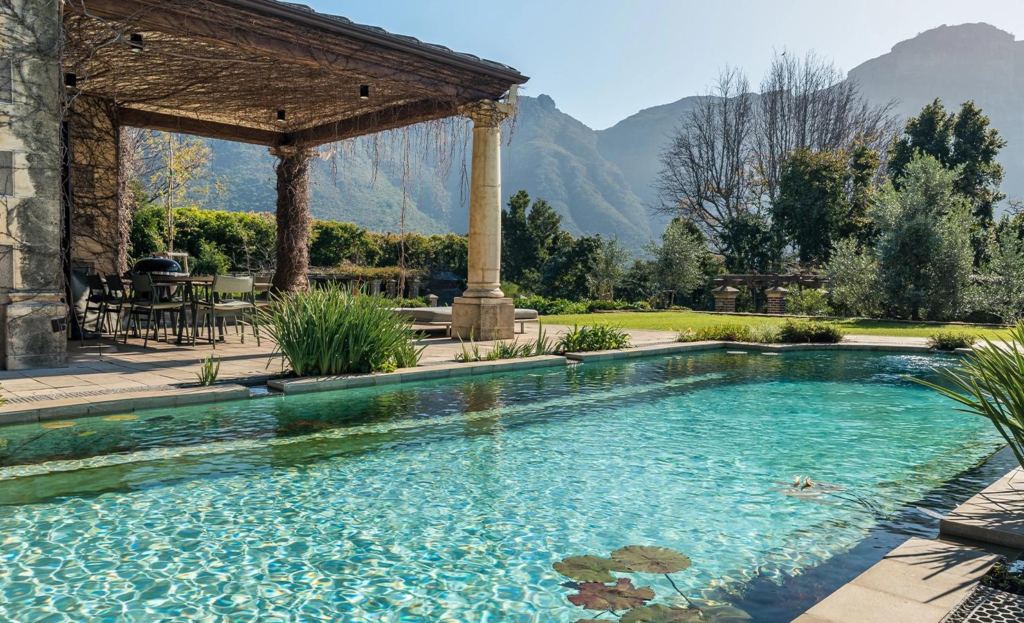 Perfect Hideaways: Natural stone-edged pool with water lilies set beside an outdoor dining pergola draped in vines, surrounded by lush gardens and magnificent mountain scenery, Bishopscourt, Bishops Hall