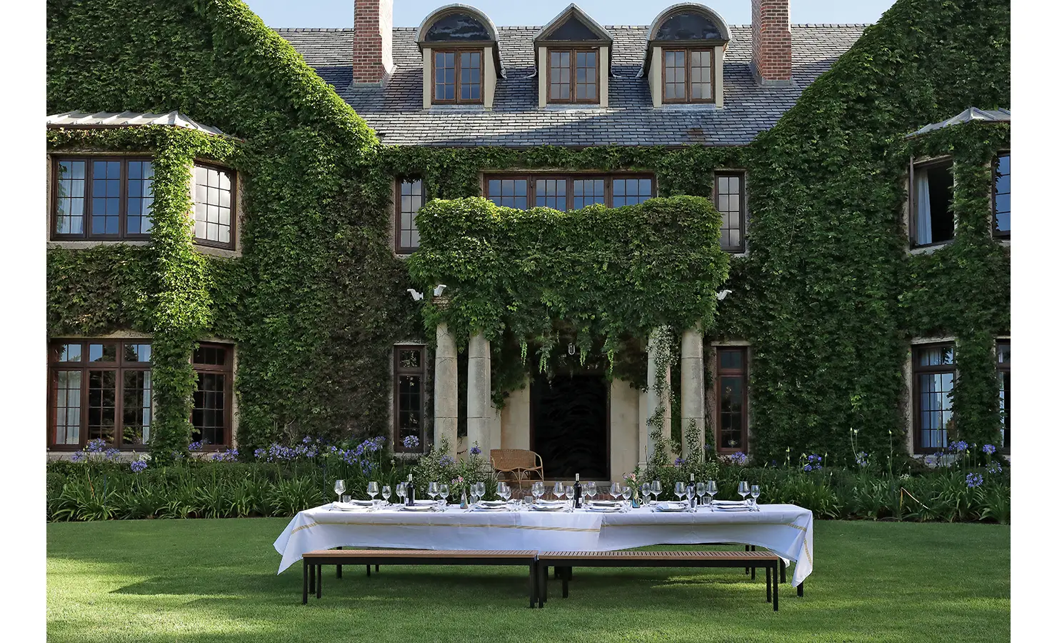 Perfect Hideaways — A long outdoor dining table elegantly set with white linens and glassware on a manicured lawn before the ivy-covered façade of a grand manor. The setting blends classic charm with understated sophistication. Bishopscourt, Bishops Hall.