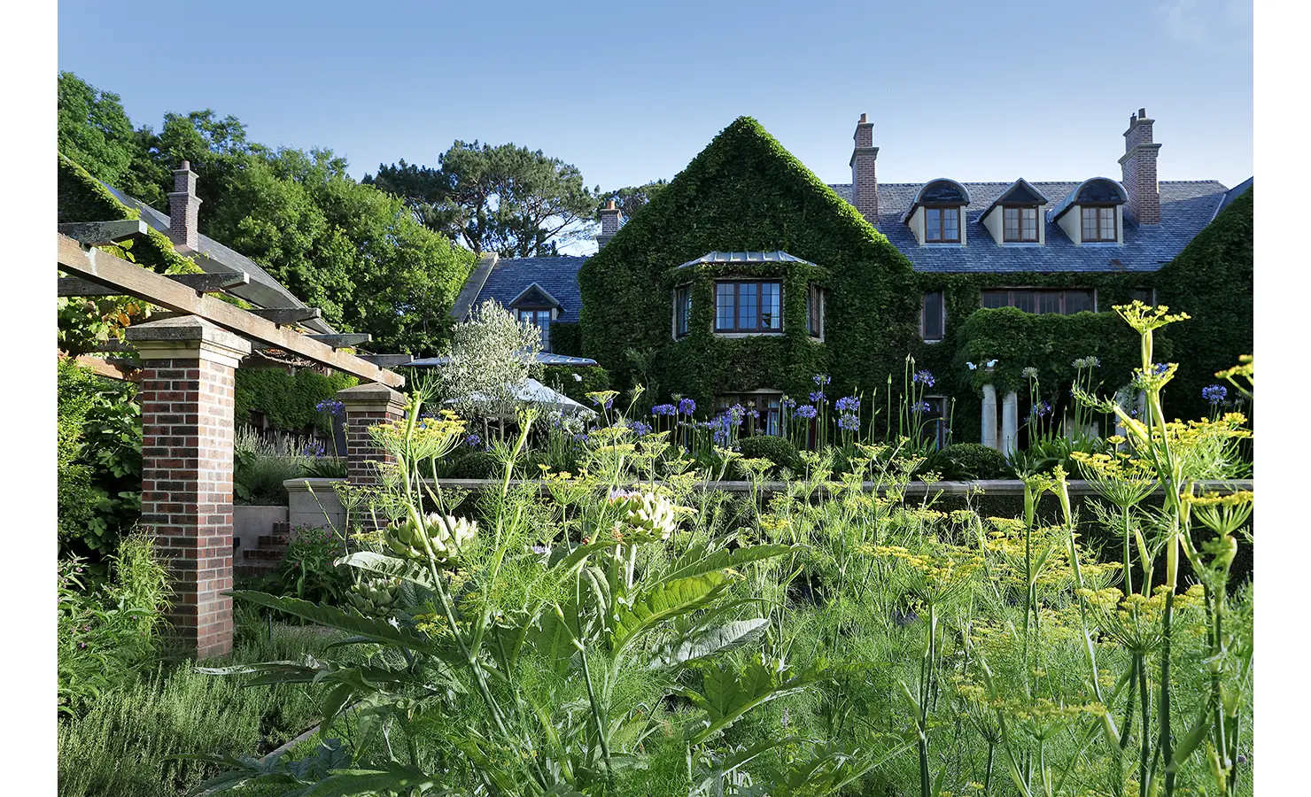 Perfect Hideaways — A view of the manor through tall garden blooms and artichoke plants, framed by brick pergolas and lush landscaping. The green façade and architectural symmetry highlight timeless elegance in nature’s embrace. Bishopscourt, Bishops Hall.