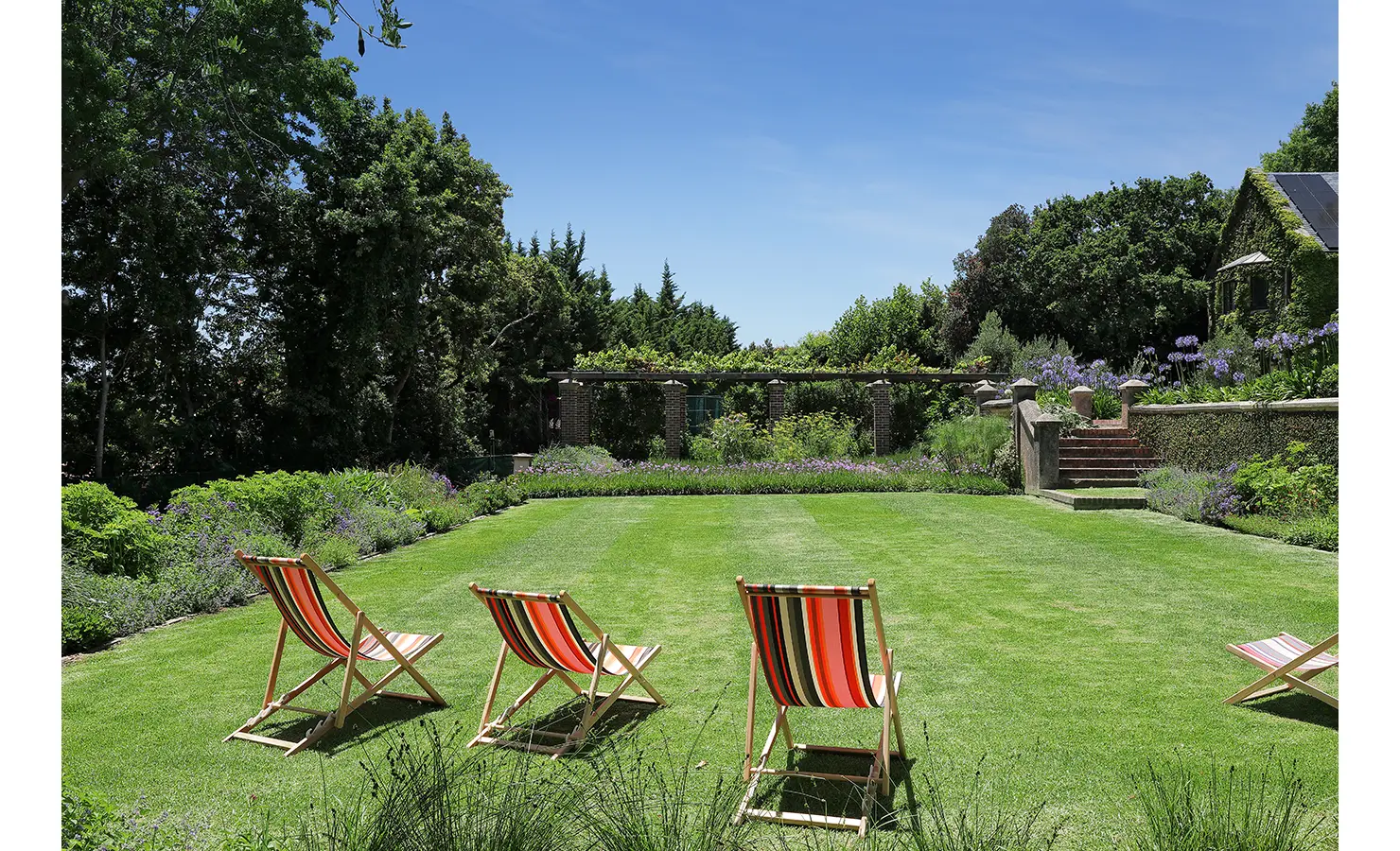 Perfect Hideaways — A perfectly kept lawn bordered by flowering hedges and shaded trees, with a row of colorful striped deck chairs inviting relaxation under the summer sun. Bishopscourt, Bishops Hall.