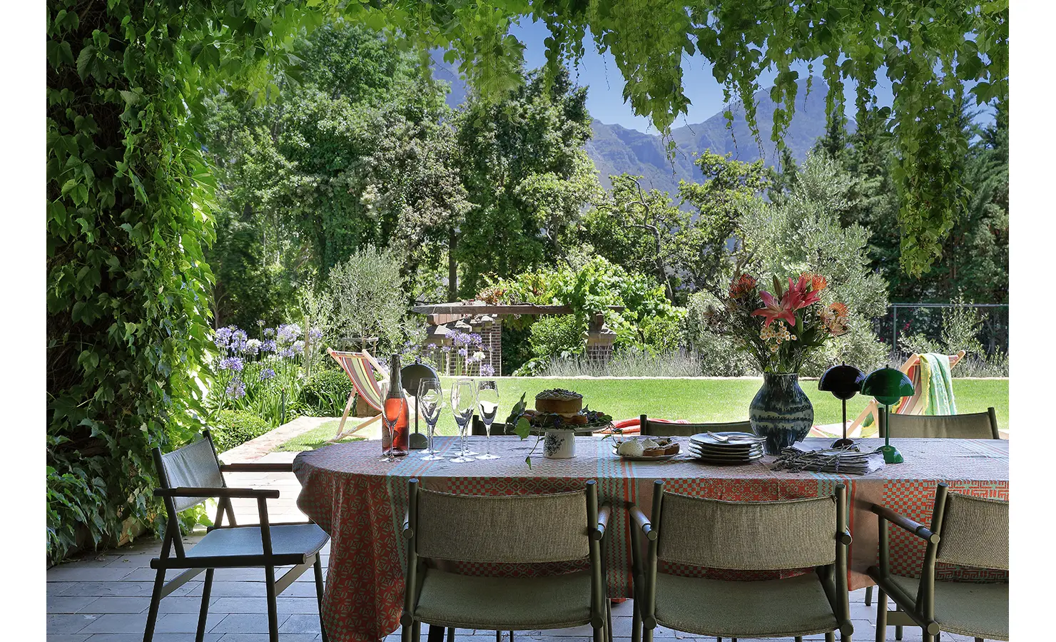 Perfect Hideaways — A shaded patio dining area draped in vines with a round table set for an al fresco meal. Fresh flowers, glassware, and garden views create a serene, inviting atmosphere with mountains in the distance. Bishopscourt, Bishops Hall.