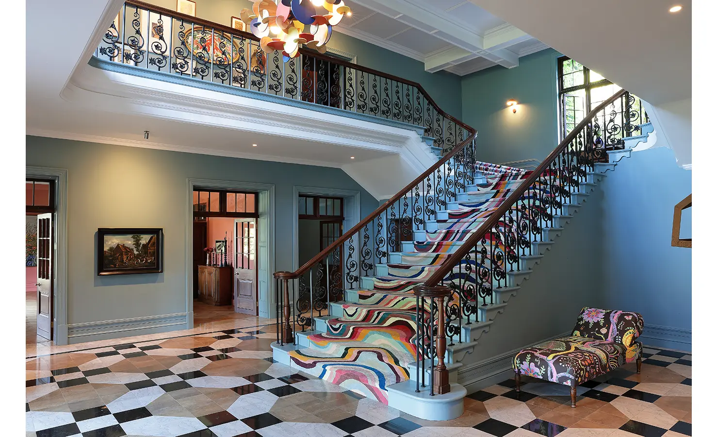 Perfect Hideaways — A grand staircase with ornate wrought-iron railings and a vibrant, multicolored runner anchors an elegant foyer. The room features checkered marble floors, modern art, and soft teal walls that exude charm and style. Bishopscourt, Bishops Hall.