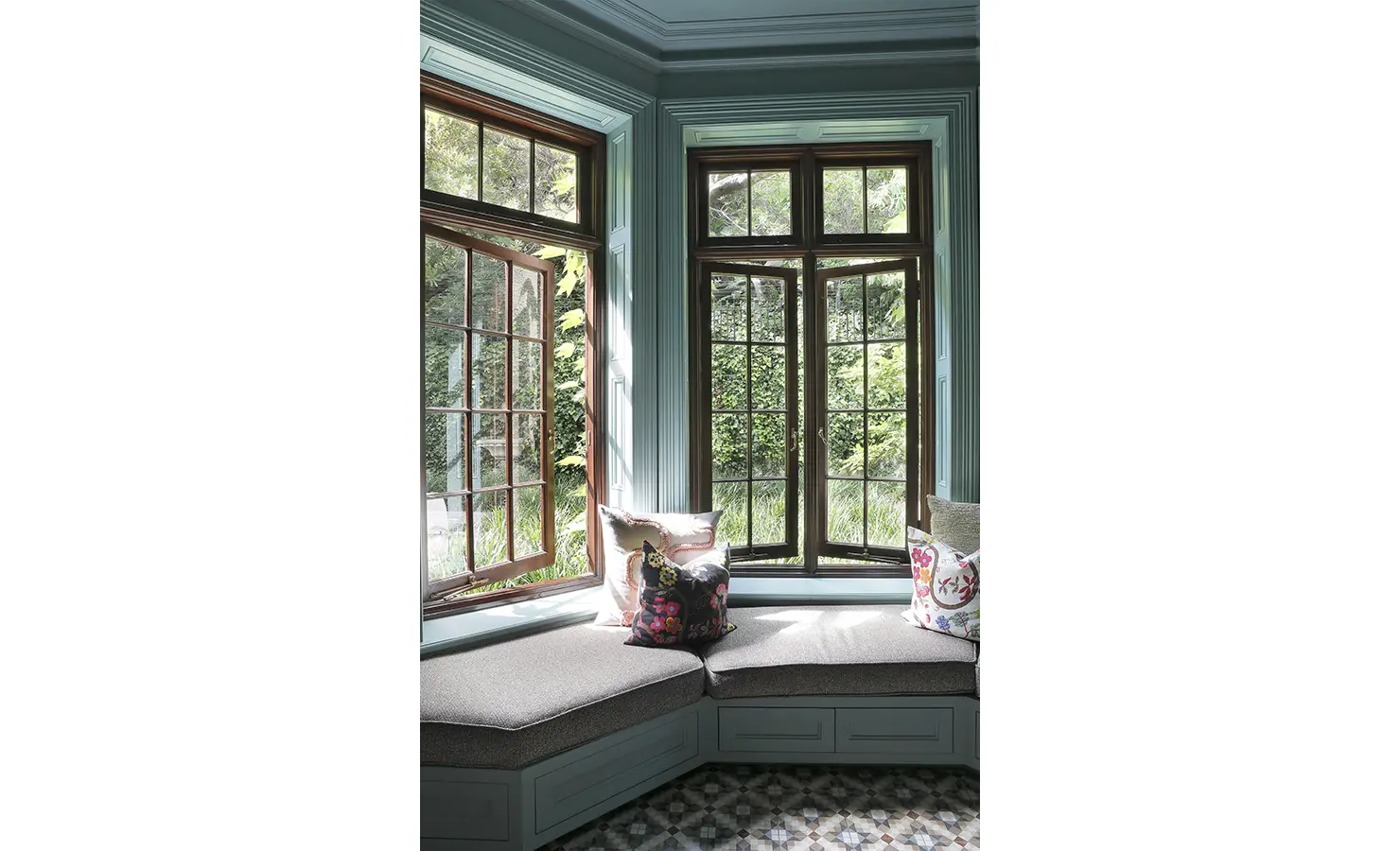 Perfect Hideaways — A cozy bay window nook with built-in seating and patterned cushions overlooks the lush garden. Sunlight filters through wooden-framed windows, illuminating the peaceful corner. Bishopscourt, Bishops Hall.